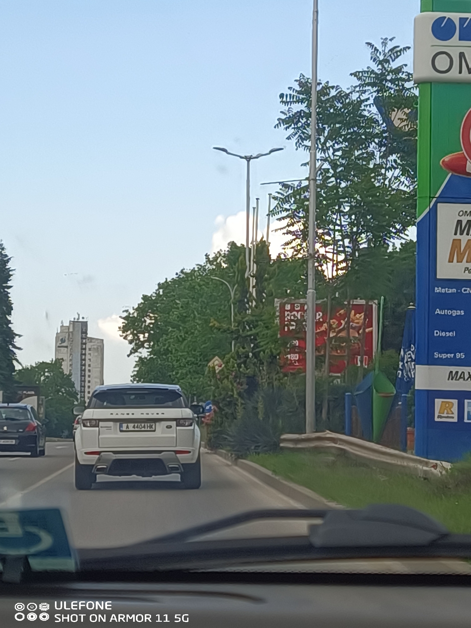 The image depicts a daytime scene from the perspective of a vehicle moving forward on an asphalt road in Burgas, Bulgaria. In the foreground, a white Range Rover Evoque with Bulgarian license plate "A 4404 HK" is observed directly ahead. A darker vehicle, possibly a Peugeot, is visible further ahead and slightly to the left.
To the right of the road, a prominent blue and green OMV gas station sign advertises "Metan-CNG," "Autogas," "Diesel," and "Super 95." Behind the OMV sign, a red billboard, partially obscured by foliage and a utility pole, displays indistinct text and the number "180." A metal guardrail and green vegetation line the roadside.
To the left of the road, dense green trees partially obscure a tall, multi-story, light-colored building with a distinct top structure, characteristic of a residential or office high-rise. The sky above is light blue with scattered white clouds. The scene is located on bul. Stefan Stambolov in Burgas, near the OMV gas station at the intersection with ul. Aleko Konstantinov, with the vehicle heading in a generally south-easterly direction towards the city center. The distant tall building is consistent with structures found in the Lazur neighborhood.
