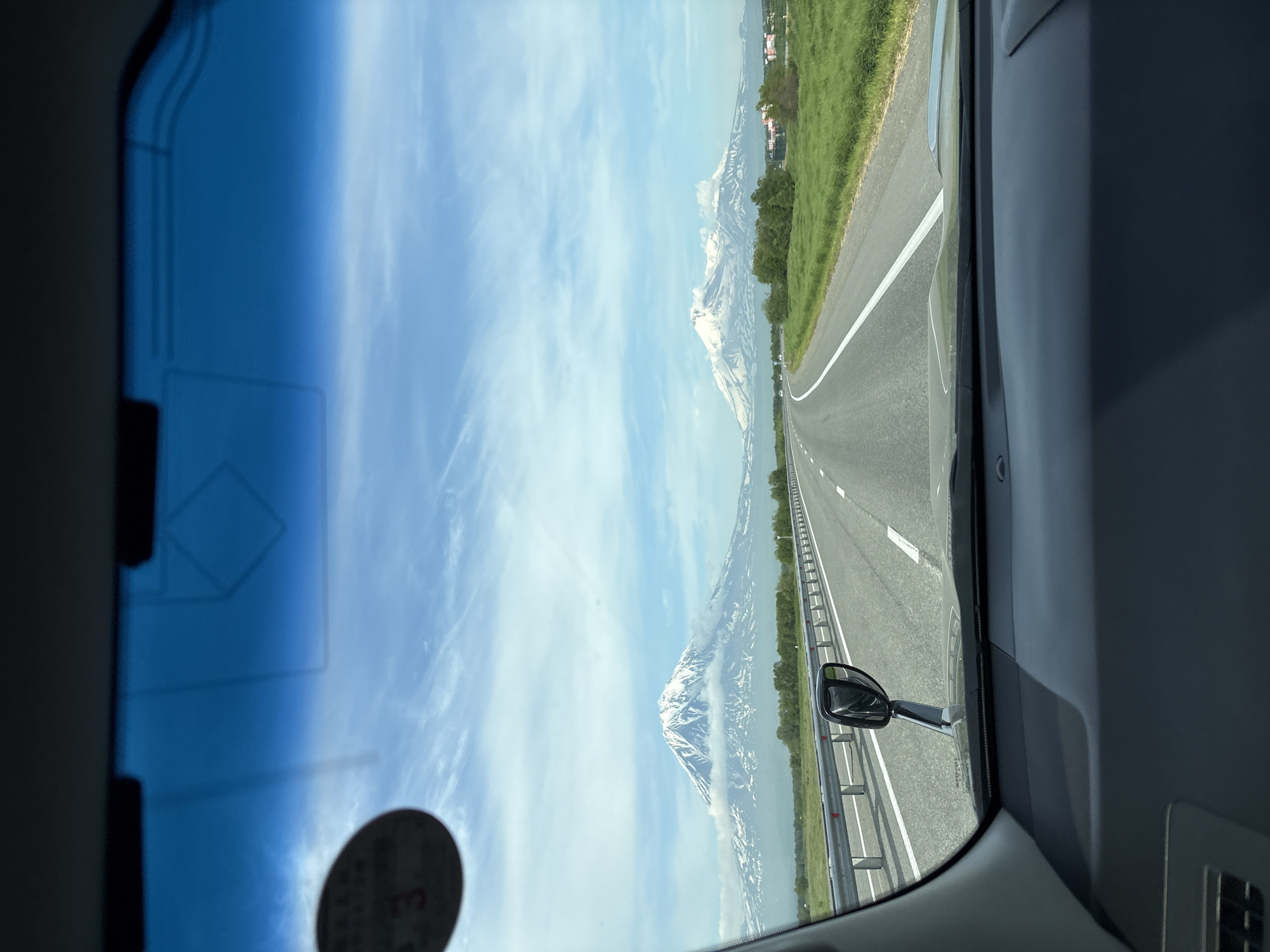 The image is a point-of-view shot from inside a vehicle, looking through the windshield. It depicts a highway leading towards two snow-capped volcanoes under a partly cloudy sky. The volcanoes are prominent features in the distance, with one larger and closer than the other. The highway is paved with white lane markings and bordered by a metal guardrail on the left. Lush green fields with scattered trees flank the right side of the road. A side mirror of the vehicle is visible on the left. The foreground shows the dashboard of the car, with some faint markings on the windshield, including a circular sticker on the upper left. The scene appears to be a scenic drive in the vicinity of Yelizovo, Russia, known for its proximity to volcanic landscapes.