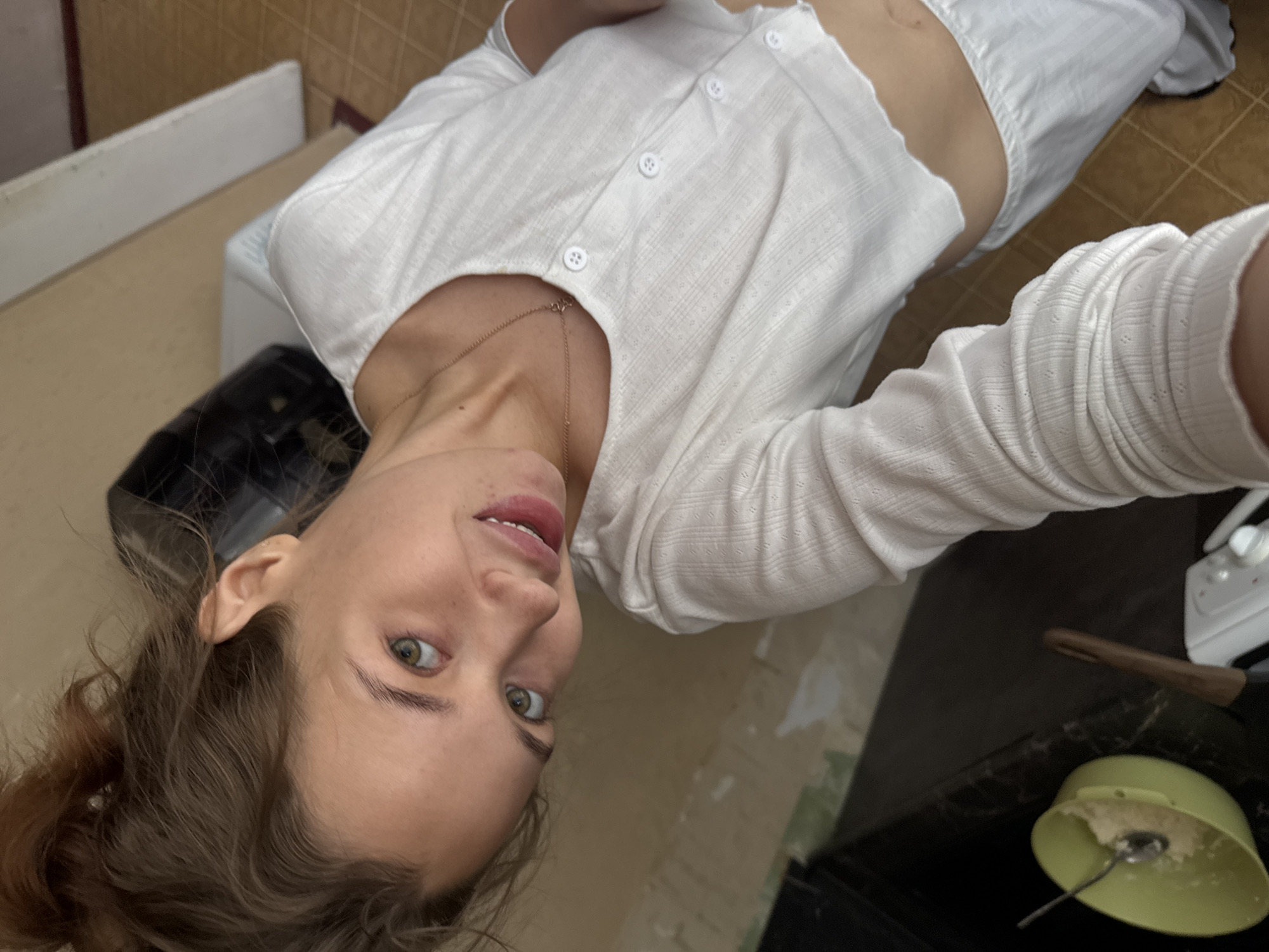 In this high-angle, close-up selfie, a young woman with light brown hair pulled up in a messy bun is shown from the chest up. She is wearing a white, cropped, long-sleeved shirt with buttons down the front and white pants. Her head is tilted, and she is looking directly at the camera with a slight smile. Her green eyes are visible, as is her collarbone and a delicate necklace.
The background suggests an indoor domestic setting, possibly a kitchen. To the left, a light green bowl containing what appears to be food and a spoon is visible on a dark countertop. A wooden-handled utensil rests beside the bowl. To the right, a white appliance with knobs is partially visible. The walls in the background show signs of unfinished renovation, with areas of exposed plaster. The floor is tiled with a checkerboard pattern of light brown and beige squares. The overall lighting is natural and diffused, indicating it may be daytime. The image is likely captured in Chaikovsky, Russia, based on the user's prompt.