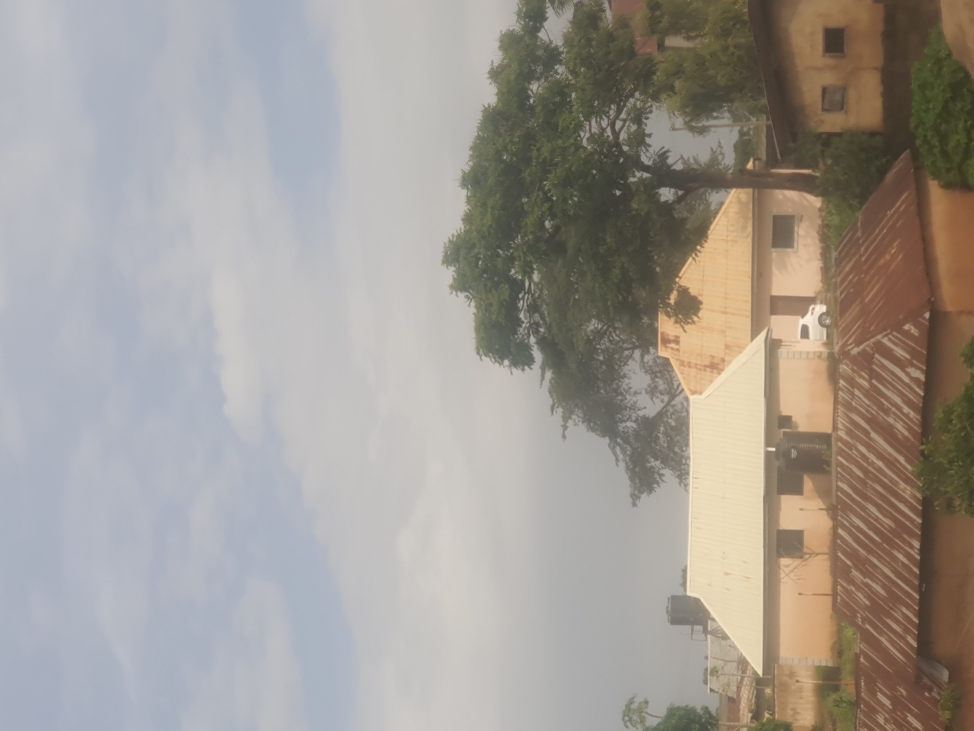 The image depicts a residential area in Jos, Nigeria, with a focus on several buildings and a large tree. The scene is viewed from an elevated perspective, looking down on rooftops and the surrounding structures.
The primary subjects are the buildings, which appear to be single-story or two-story houses with varying roof types. Some roofs are made of corrugated metal, showing signs of rust, while others are lighter colored, possibly metal or a coated material. One building has a cream-colored, peaked roof. Visible architectural details include windows, some of which are dark squares suggesting they are covered or unlit, and others are open or lighter. One building has water tanks on its roof.
A prominent feature is a large, leafy tree with dense foliage, casting shadows on the buildings and landscape. Its branches extend across a significant portion of the frame.
A white car is parked in front of one of the buildings, partially obscured by the tree.
The sky is overcast with some visible clouds, suggesting a diffused light. The overall atmosphere is somewhat hazy. The time of day appears to be daytime, likely late morning or afternoon, given the lighting. The environment suggests a relatively dense urban or semi-urban setting, common in residential areas of Jos.
There is no visible text in the image. The image captures a still moment of everyday life in a Nigerian residential area.