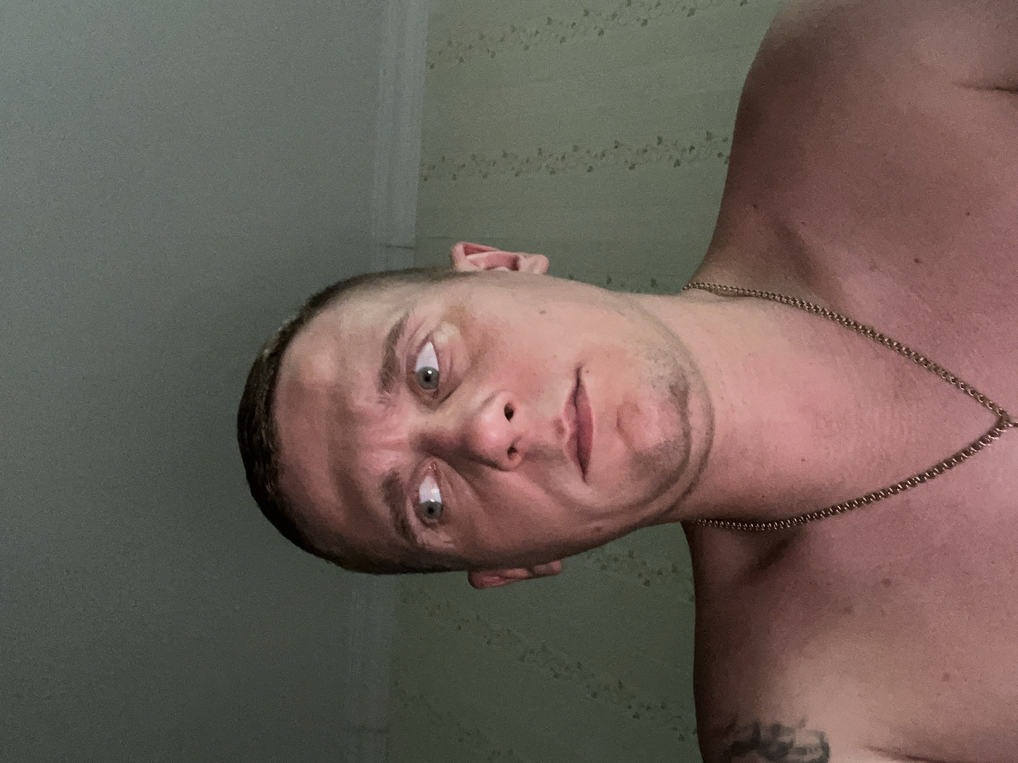 A close-up image depicts an adult male from the chest up, positioned indoors. The man has short, dark hair and light-colored eyes. He is bare-chested, wearing a gold-colored chain necklace with a pendant, and a dark tattoo is partially visible on his left shoulder. His head is turned slightly, and his eyes are wide, focused intently towards the left of the frame. His lips are closed and slightly pursed, suggesting a focused or strained expression. The background features light green/teal walls with a faint, darker, repeating floral or vine-like pattern, and a light-colored ceiling line is visible at the top. The lighting appears to originate from above, casting subtle shadows on his face. The indoor setting and wall patterns are consistent with a residential or private space. The image provides no visual cues or identifiable landmarks to confirm the specific location as Minsk, Belarus.