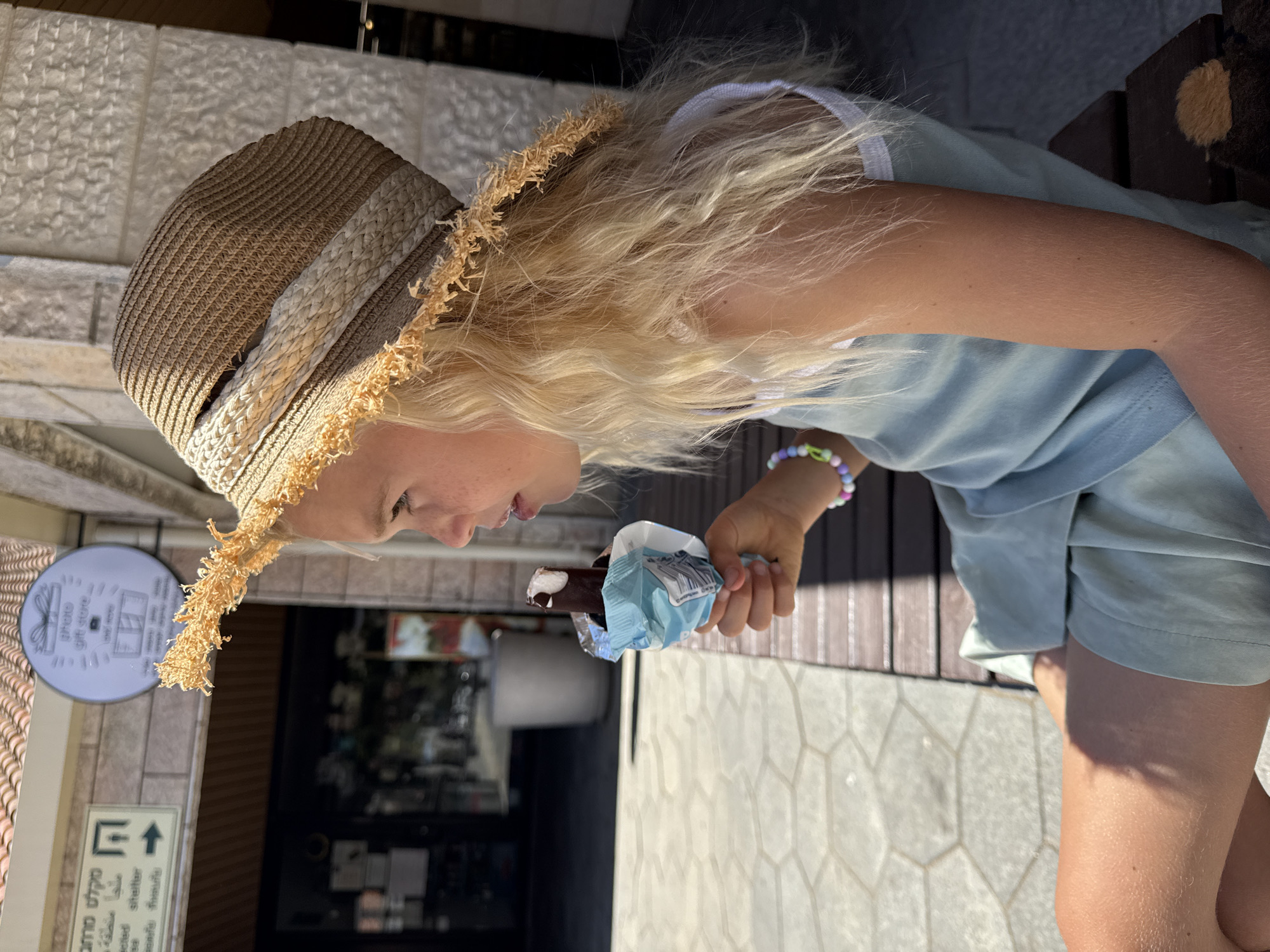 A young blonde girl is sitting on a wooden bench outside a shop in Kfar Vradim, Israel, eating an ice cream bar. She is wearing a straw hat with frayed edges and a light blue dress. The shop behind her has a sign with Hebrew writing and a circular logo. The pavement in front of the bench is made of hexagonal tiles. The lighting suggests it is a sunny day.