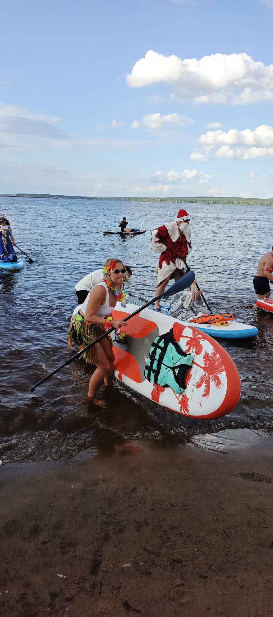 In Chaikovsky, Russia, on a sunny day with scattered clouds, a group of people are stand-up paddleboarding on a wide body of water, likely a river or lake, with a tree-lined shore in the distance. In the foreground, a woman wearing sunglasses, a white tank top, and a colorful grass skirt is standing in the shallow water, holding a paddle and positioned on an orange and white paddleboard decorated with palm trees. To her right, a person dressed as Santa Claus with a white beard and red suit is standing on a paddleboard, holding a paddle. Further back, another paddleboarder is seen wearing a blue and white traditional dress, and in the mid-ground, another individual is lying on a paddleboard. The water is calm and appears to be near a muddy shore.