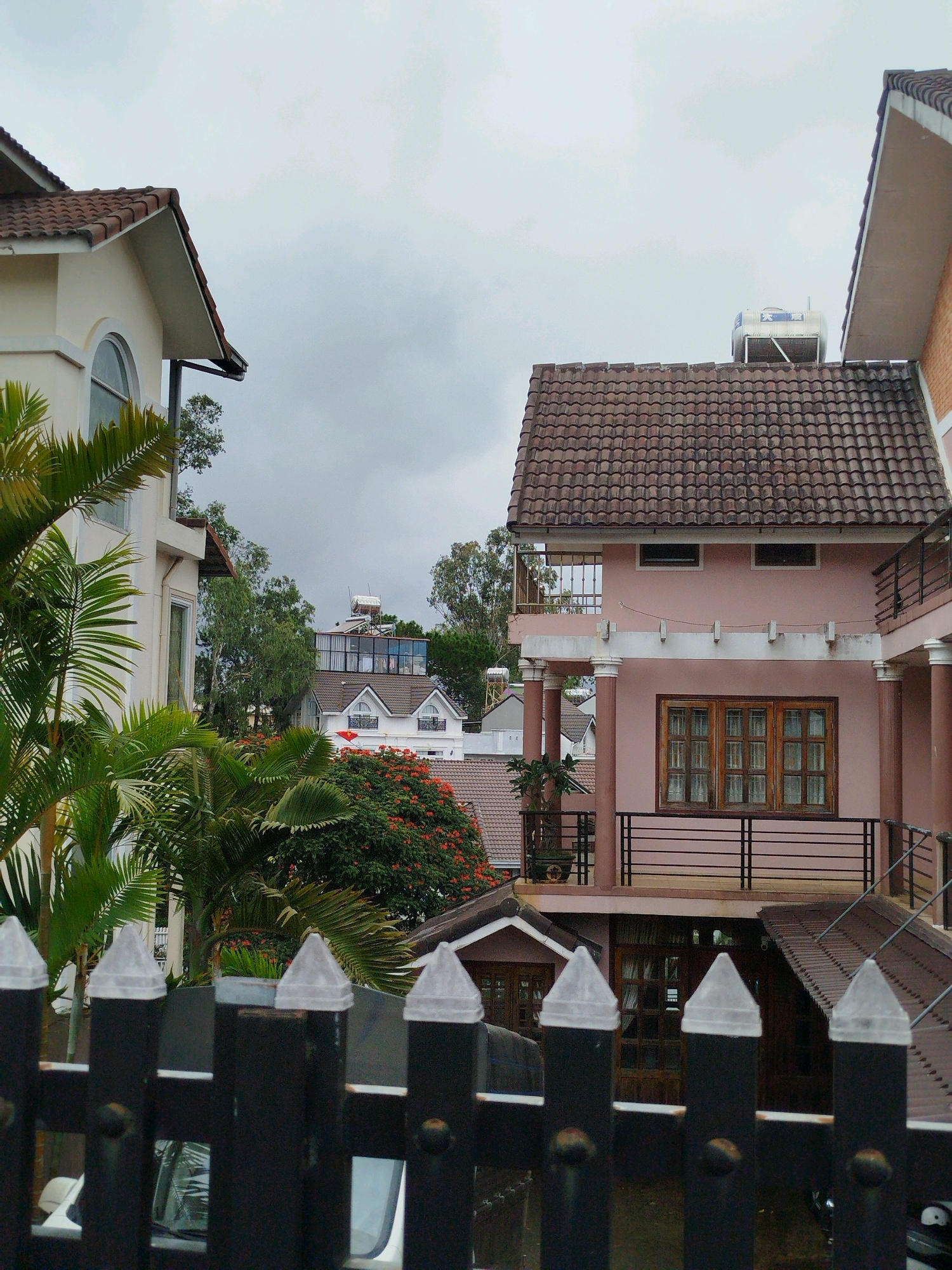 The image depicts a view of buildings with tiled roofs and balconies, set against a cloudy sky. In the foreground, a black metal fence with pointed translucent caps is visible, partially obscuring a car. Lush green palm fronds and a tree with red flowers frame the scene on the left and in the middle ground, respectively. Several houses are visible, one painted pink with wooden-framed windows, another a lighter color with arched windows, and a white multi-story house with a Vietnamese flag prominently displayed. On the roofs of some buildings, water tanks and solar water heaters are present. The architectural style suggests a residential area, possibly in a city like Da Lat, Vietnam, known for its French colonial-inspired villas and cooler climate, indicated by the lush vegetation and tiled roofs designed to handle rain. The overall atmosphere is overcast, suggesting it might be a cloudy or rainy day.