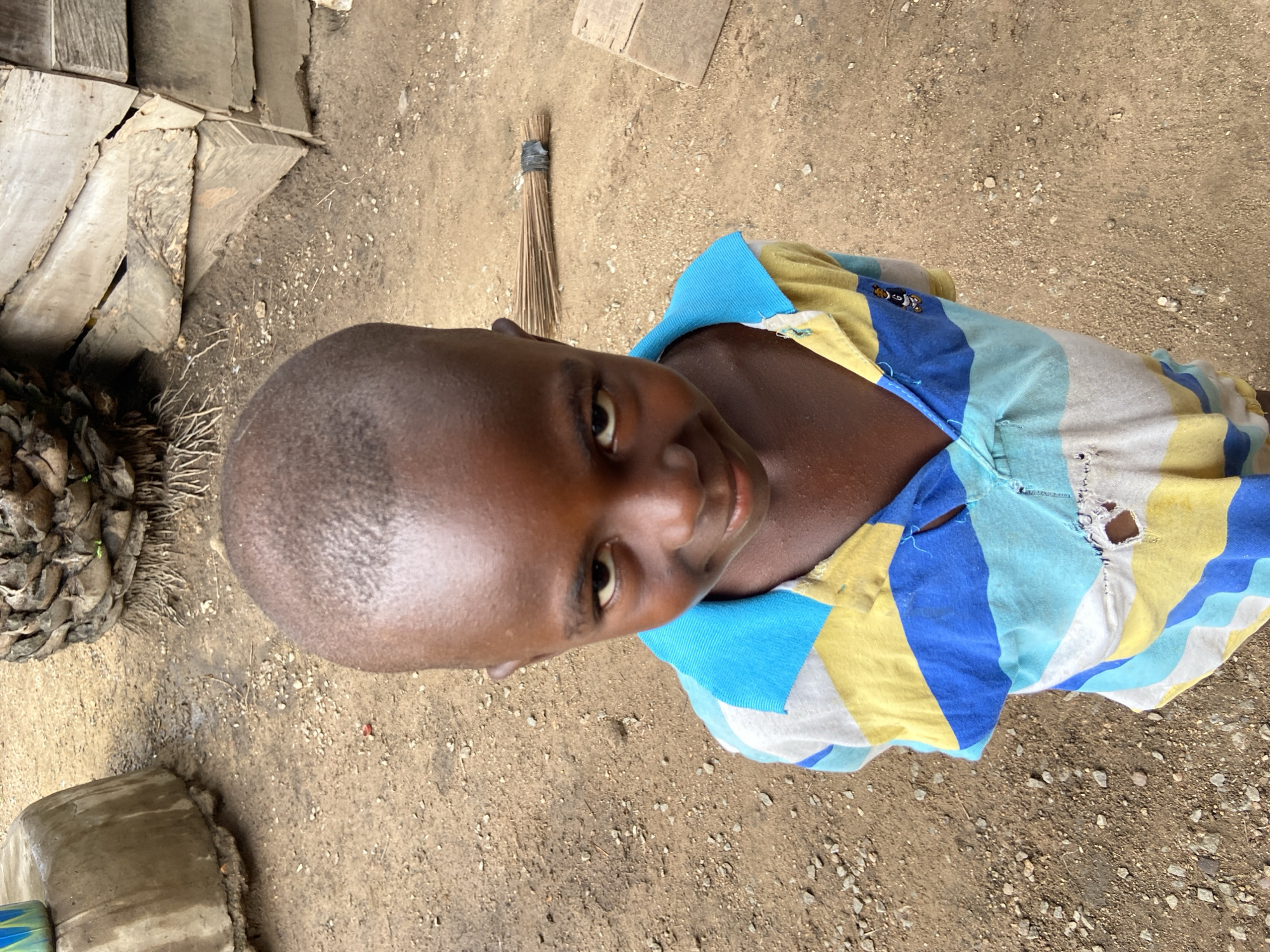 A boy stands on a dirt ground surface with pebbles. He wears a blue and yellow striped shirt with a hole. Beside the boy is a broom, a concrete container, wooden planks, and a dried flower arrangement. The location is Mararaba, Nigeria.