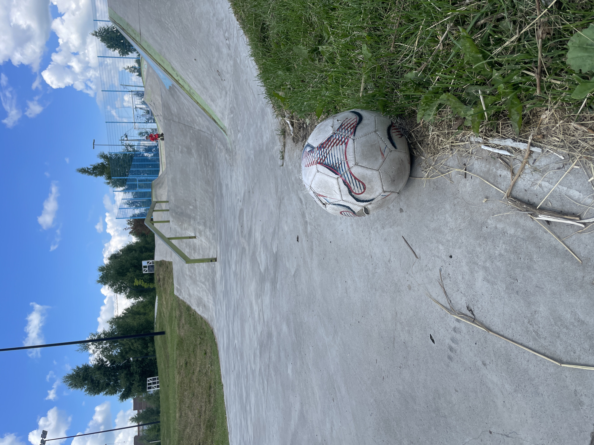 An outdoor shot of a skatepark in Laishevo, Russia, under a partly cloudy sky. The foreground is dominated by a worn soccer ball lying on a concrete surface next to a patch of grass. Beyond this, the skatepark features concrete ramps, rails, and a blue fence. In the distance, trees and buildings are visible. A child on a scooter is seen in the mid-ground. The scene appears to be set during daylight hours.