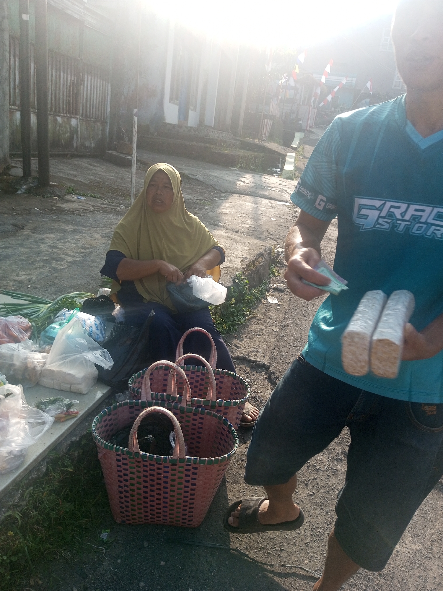 In Krasak, Indonesia, an outdoor scene depicts a woman wearing a hijab and a headscarf, seated on the ground near a collection of plastic bags filled with groceries. She appears to be engaged in a transaction, holding a plastic bag. In the foreground, a person in a blue t-shirt and denim shorts is shown holding cash and two food items wrapped in plastic. The background features a building with a corrugated metal fence and a street with a light-colored pavement. Sunlight streams in from the right side of the image, creating a bright and airy atmosphere. Several woven baskets are also visible in the foreground.