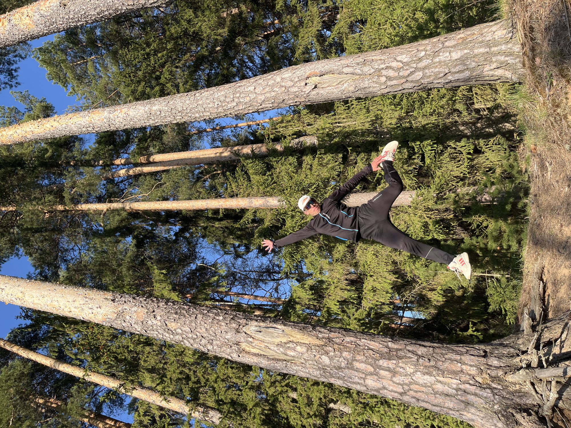 The image shows a person in a forest, appearing to be performing a horizontal split between two trees. The individual is wearing a black athletic outfit with a white baseball cap and white sneakers. The background consists of tall pine trees and a clear blue sky visible through the canopy. The location is assumed to be in a forest or park area, possibly in or near the city of Chaikovsky, Russia. The person is positioned horizontally, with one foot resting on one tree trunk and the other foot resting on another tree trunk, attempting a split.