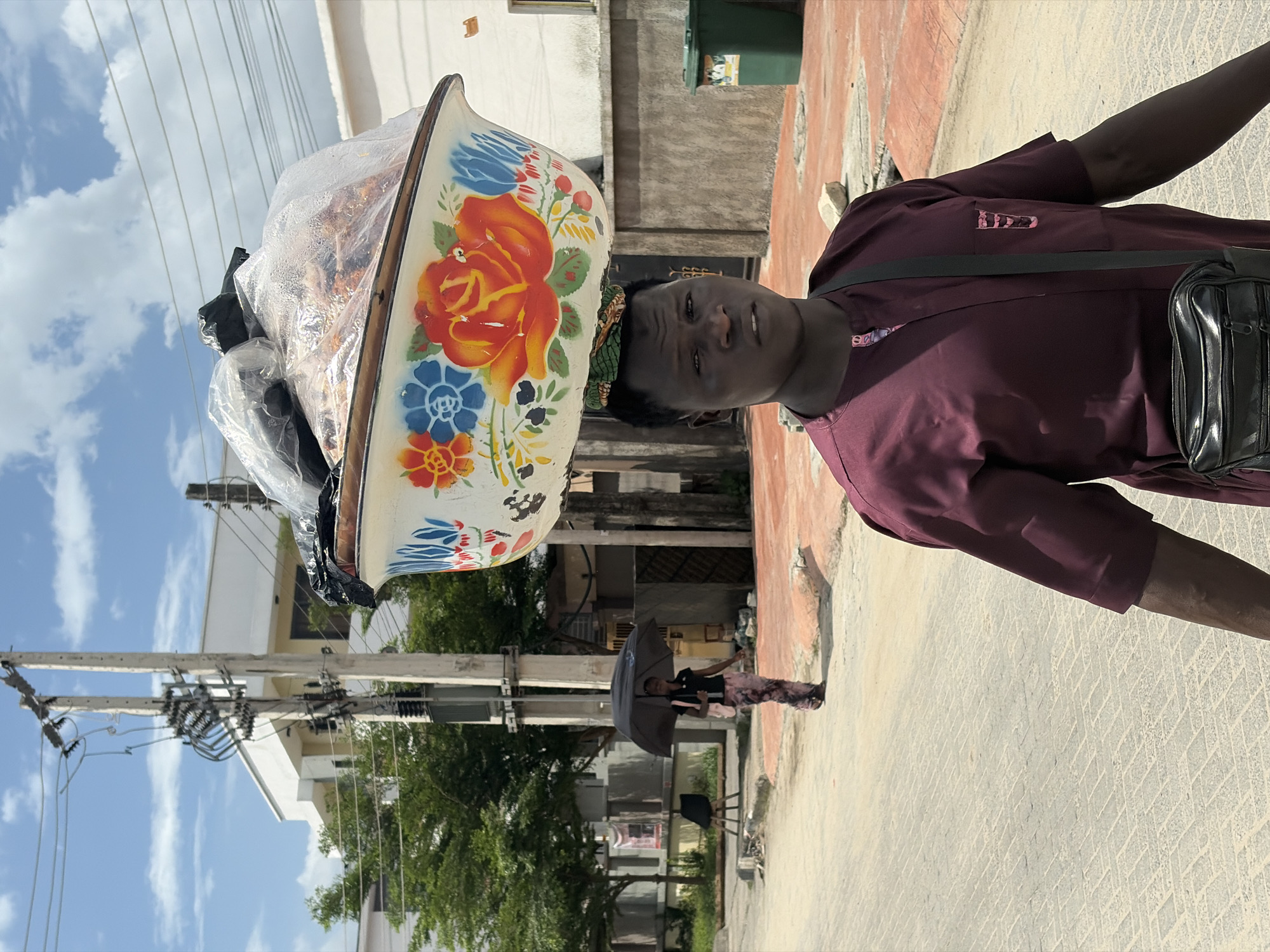 A man in a maroon shirt with a black shoulder bag carries a large, round, floral-patterned bowl filled with food on his head, while standing on a paved street in Igboefon, Nigeria. Another person holding an umbrella is visible in the background. Utility poles with electrical wires are on the left. Buildings and trees line the street. The sky is visible in the upper left.