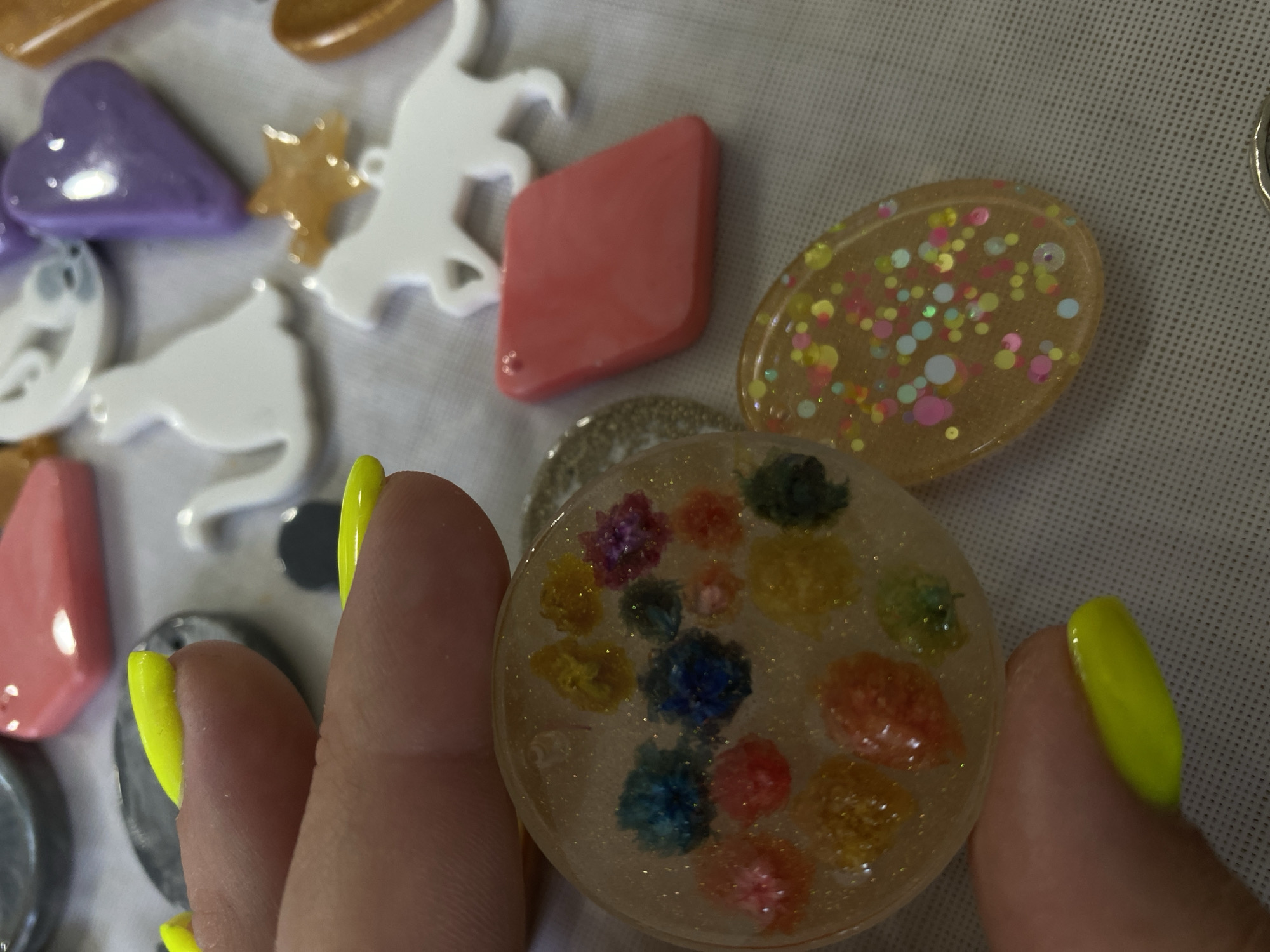 A close-up shot captures a person's hand with bright yellow fingernails gently holding a circular pendant. The pendant is clear resin filled with tiny, colorful dried flowers and shimmering glitter. In the background, a collection of similar decorative charms are scattered on a white textured surface. These charms include various shapes like cats, horses, hearts, stars, and geometric forms, rendered in different materials and colors such as white, purple, pink, orange, and grey. The scene is devoid of any specific urban or geographical markers, making it impossible to determine a location within Balashikha, Russia, or any other city.