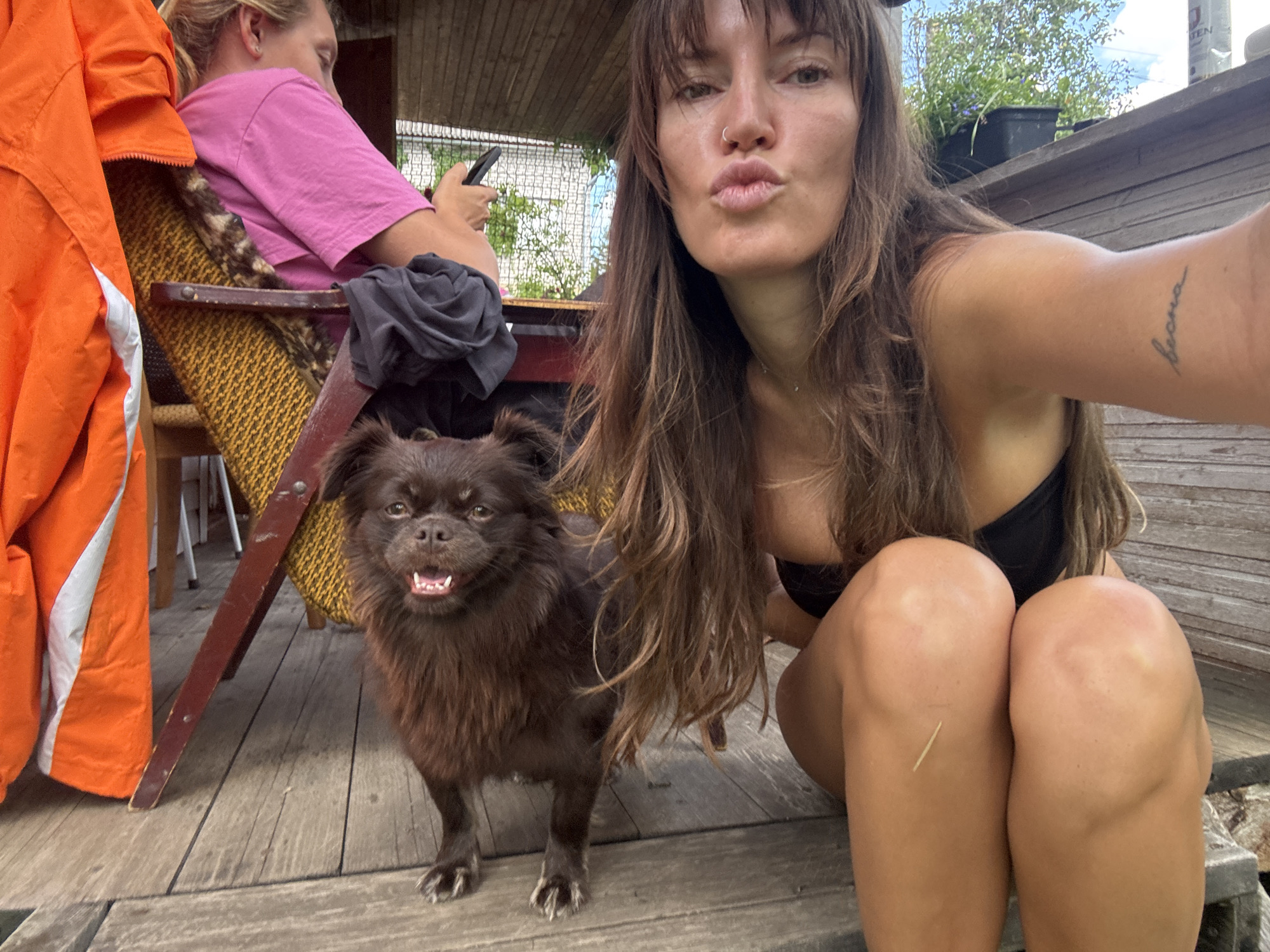 In a low-angle, outdoor, medium shot, a young woman with long brown hair and bangs is captured in a selfie. She is wearing a black bikini top and has a nose ring. Her mouth is pursed in a kissing gesture. To her left, a small, fluffy brown dog stands on a wooden deck, its mouth open and tongue visible, appearing to be panting or smiling. Behind the woman, another person in a pink t-shirt is partially visible, sitting in a chair and looking at a mobile phone. An orange jacket hangs on the left side of the frame. The background features a wooden structure with a partial roof, greenery, and a white railing. The scene appears to be an outdoor setting, possibly a backyard or a dock, in Красная Пресня Снт, Russia.