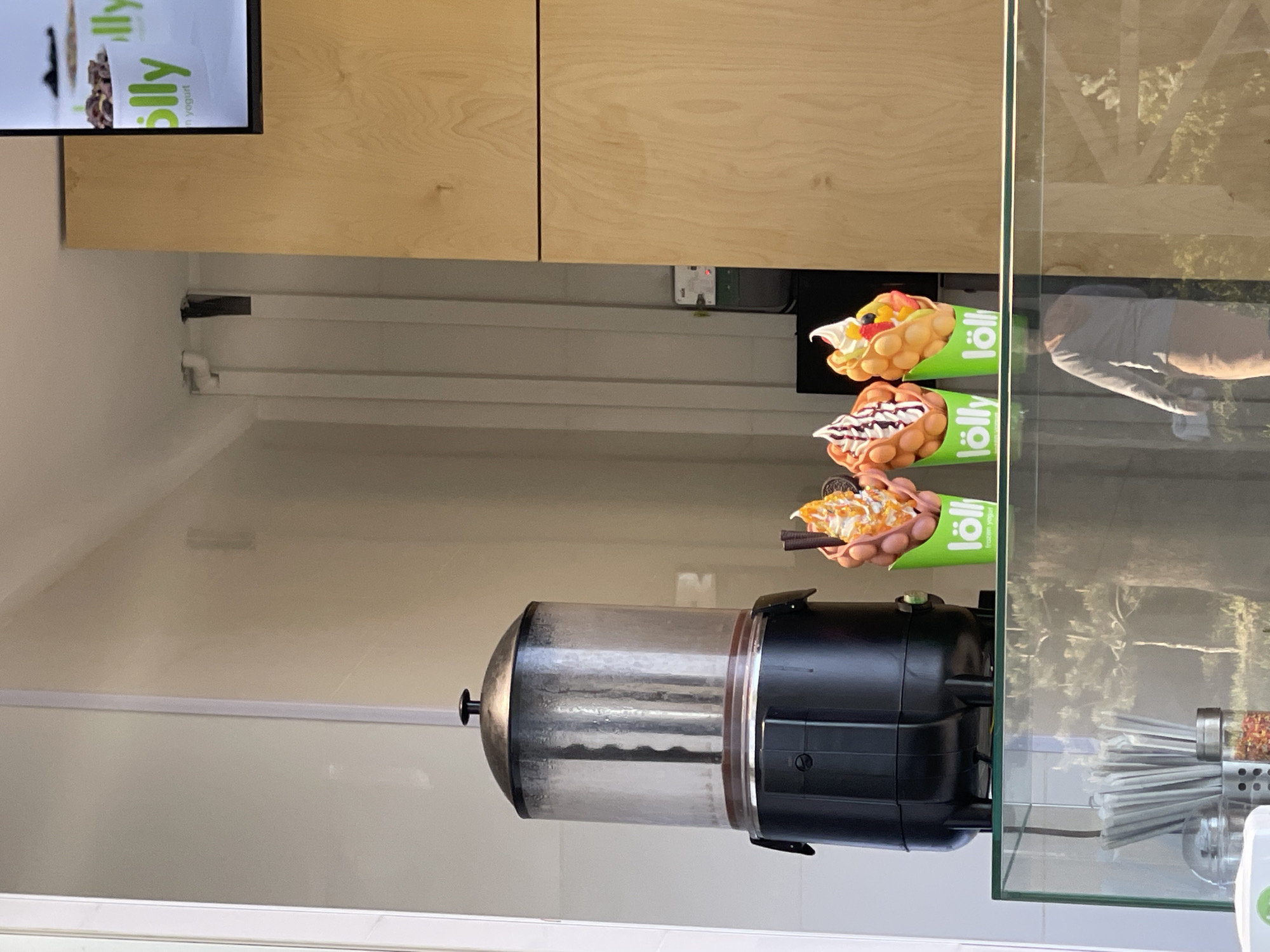 This image, captured inside a food establishment in Tbilisi, Georgia, showcases a display counter with various dessert offerings and equipment. The scene is lit by indoor lighting, making it difficult to determine the time of day.
In the upper left portion of the frame, a digital screen displays text. The prominent brand name "lölly" is visible in green, along with the word "yoghurt."
Below the screen and extending across the middle of the image are light wooden cabinets or wall panels, suggesting a clean and modern aesthetic.
To the right of the wooden panels, a row of three distinct dessert items is displayed, each presented in a conical, bright green holder also branded with "lölly." These appear to be Hong Kong-style egg waffles (bubble waffles) with various toppings:
* The uppermost waffle is generously topped with an assortment of fresh fruit pieces and white whipped cream.
* The middle waffle features a dark chocolate drizzle over white whipped cream.
* The lowermost waffle is adorned with cream and what appear to be crushed cookies or similar toppings, with a pair of chopsticks partially holding it.
In the lower-right foreground, a large beverage dispenser is visible. It has a transparent cylindrical upper section, likely for holding a drink like hot chocolate or juice, resting on a black base. Next to it, on the far right, a vertical glass display case reflects parts of the interior and a blurry reflection of a person (torso and arm) as well as some outdoor foliage or structures. Inside this glass case, various straws and possibly condiment shakers are arranged.
The background consists of light-colored walls with some visible conduits or wiring. The overall impression is that of a contemporary dessert or snack shop ready to serve customers.