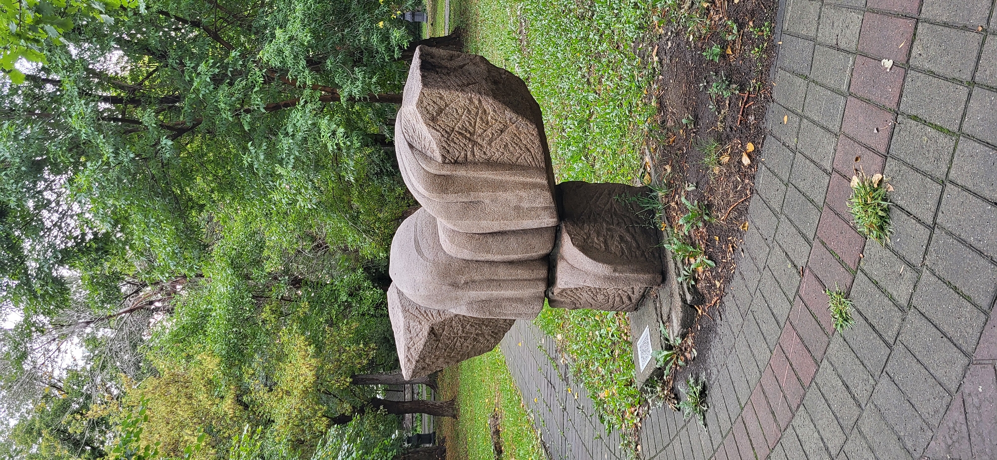 The image shows a large, abstract stone sculpture situated in a park-like setting. The sculpture is composed of several rough-hewn, angular stone blocks that are stacked and balanced to create a sense of dynamic tension. The stone has a rough, textured surface with visible tool marks and some areas of erosion. It is light brown or tan in color. The sculpture is mounted on a base of smaller stone blocks. In front of the sculpture, on the paved walkway, is a small white square object, possibly a QR code or a plaque. The walkway is made of interlocking rectangular pavers, with some areas of red and grey bricks laid in a herringbone pattern. The ground surrounding the sculpture is covered with green grass, dirt, fallen leaves, and some weeds. In the background, there are numerous trees with dense green foliage, suggesting a temperate climate and possibly autumn. The sky is overcast and pale. The overall impression is of an outdoor public art installation in a natural environment. The sculpture is located in the city of Oskemen, Kazakhstan.