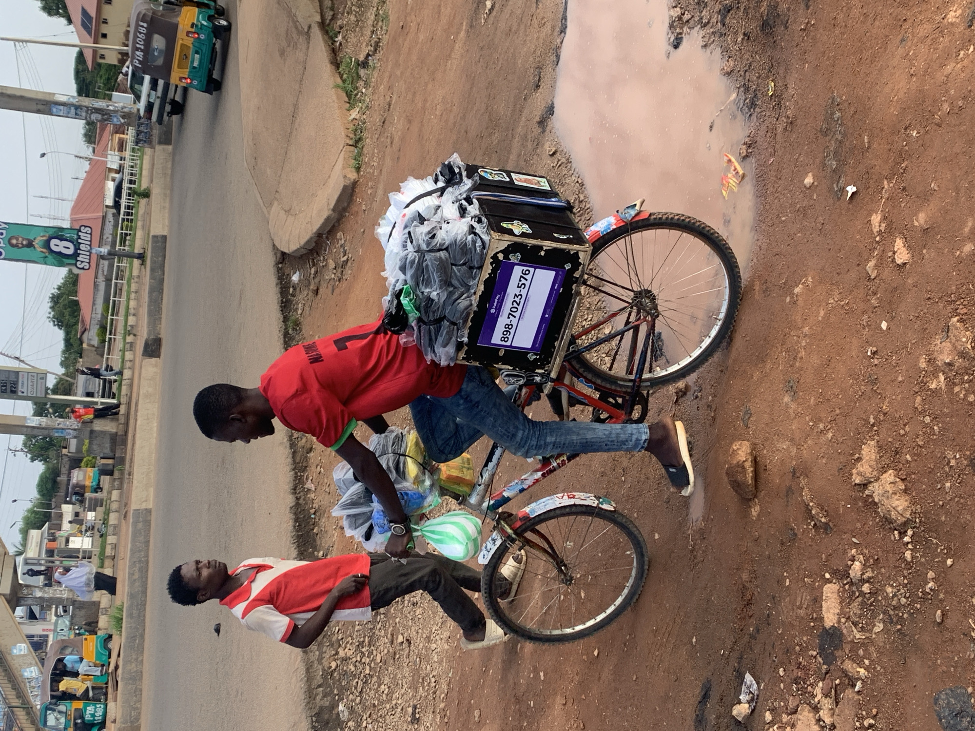 This image depicts an outdoor scene on a dirt road, likely in Gura Top, Nigeria. Two young men are present. One is standing and appears to be conversing with the other, who is seated on a bicycle. The man on the bicycle is wearing a red t-shirt and blue jeans, and he is carrying a large quantity of items, mostly in plastic bags, on the back of his bicycle in a wooden crate. The crate has a purple sign with white lettering that reads "PayKee" and a phone number "898-7023-576". The man standing is wearing a red and white polo shirt and dark pants. The environment is characterized by a dirt road with several puddles, suggesting recent rain. In the background, a street with vehicles, including a yellow and green tricycle, and buildings are visible. There is also a large billboard featuring an image of a man and text. The overall impression is one of a bustling street scene in a Nigerian city, with a vendor preparing to move his goods.