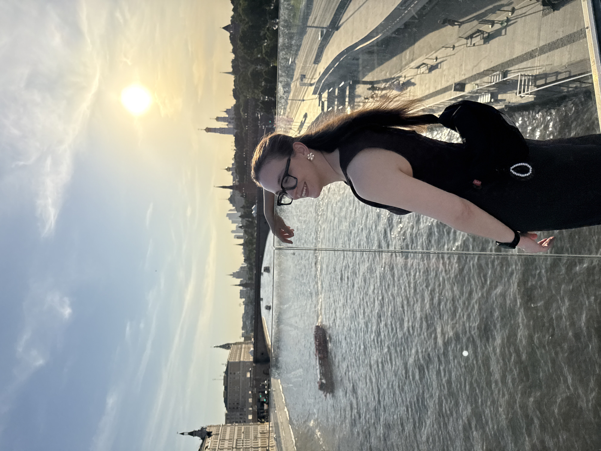 A woman stands on the Zaryadye Park Overpass in Moscow, Russia, overlooking the Moskva River. She wears a black dress, glasses, and a black backpack. The sun is visible in the sky. Buildings line the riverbank in the background.