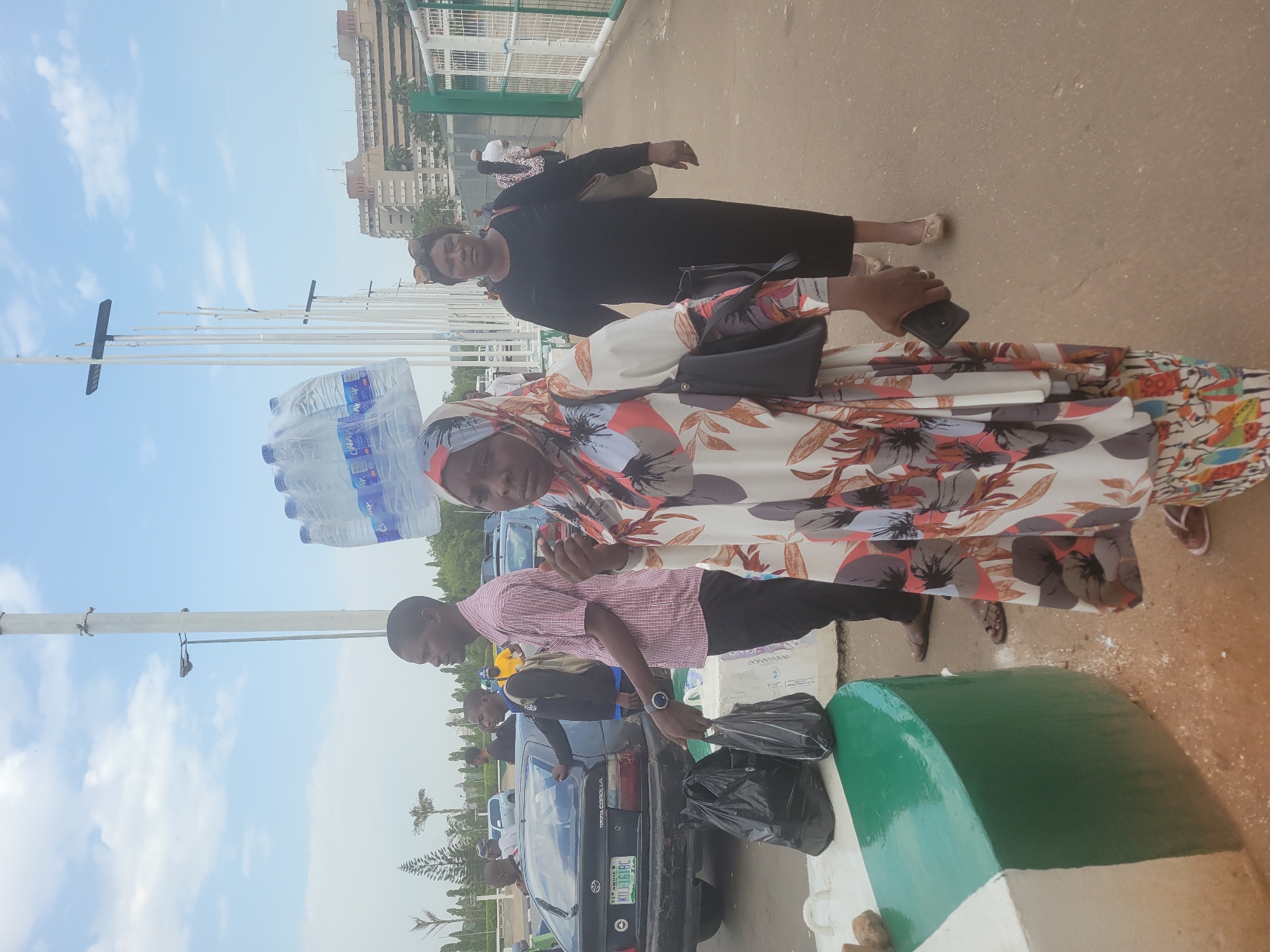 The image shows a group of people on what appears to be a sidewalk or pedestrian area, likely located in Abuja, Nigeria.
In the foreground, a man is placing a black plastic bag into the trunk of a black Toyota Corolla car. The car has a Nigerian license plate. Beside him, a woman wearing a floral patterned dress and head covering is holding a stack of bottled water. Another woman in a black dress stands nearby. The sky is visible with light clouds. Tall white flagpoles are in the background. A building is also visible in the distance. The overall scene appears to be an outdoor gathering or outing.