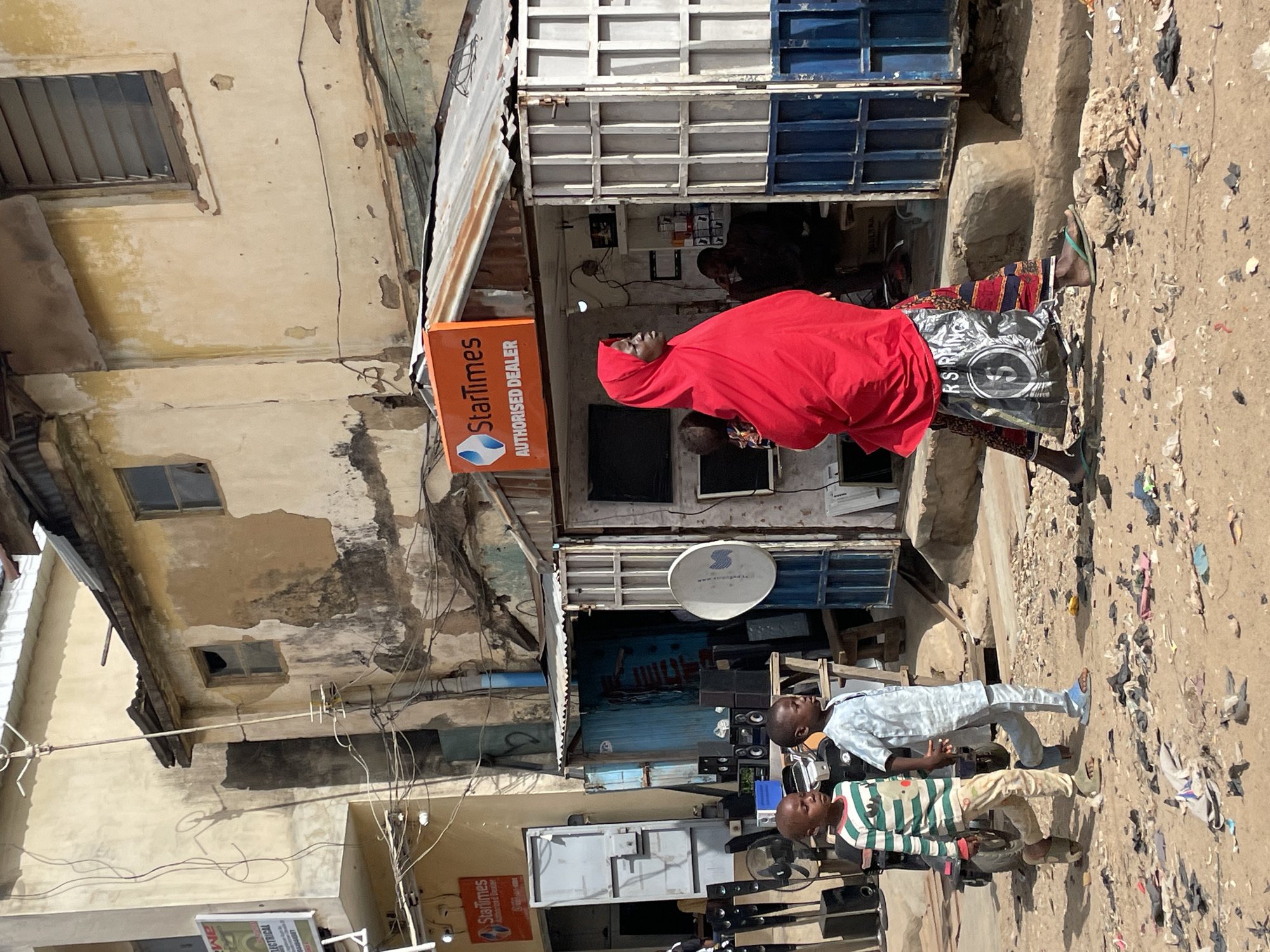The image captures an upright street scene in Jos, Nigeria, under bright daylight, suggested by strong shadows. The setting appears to be an urban commercial area with shops and residential buildings lining a dirt ground.
In the mid-ground, a woman in a vivid red head covering and top, paired with a patterned skirt and flip-flops, stands near the entrance of a shop. She is facing towards the left side of the frame. Attached to the building next to her is a prominent orange sign with white text, which reads: "StarTimes AUTHORISED DEALER". The shop entrance features blue and white shutter-style doors, and various electronic items like speakers and screens are displayed outside and inside. A person can be vaguely seen inside the shop. Above the shop, a white satellite dish is visible.
In the foreground, two young boys stand on the sandy ground, which is littered with debris including plastic, paper, and fabric scraps. One boy wears a light blue long-sleeved tunic and light trousers with light blue sandals. The other boy wears a horizontally striped green, white, and beige long-sleeved top with light-colored trousers and darker sandals. They appear to be observing the surroundings or the items displayed.
The background shows aged buildings with peeling light-colored paint, exposed concrete, and multiple windows, some with louvers. Numerous electrical wires crisscross between buildings, indicating a dense urban environment. Corrugated metal sheets are used for roofing or awnings on some structures. The overall atmosphere suggests a busy, weathered, yet active market or residential street.