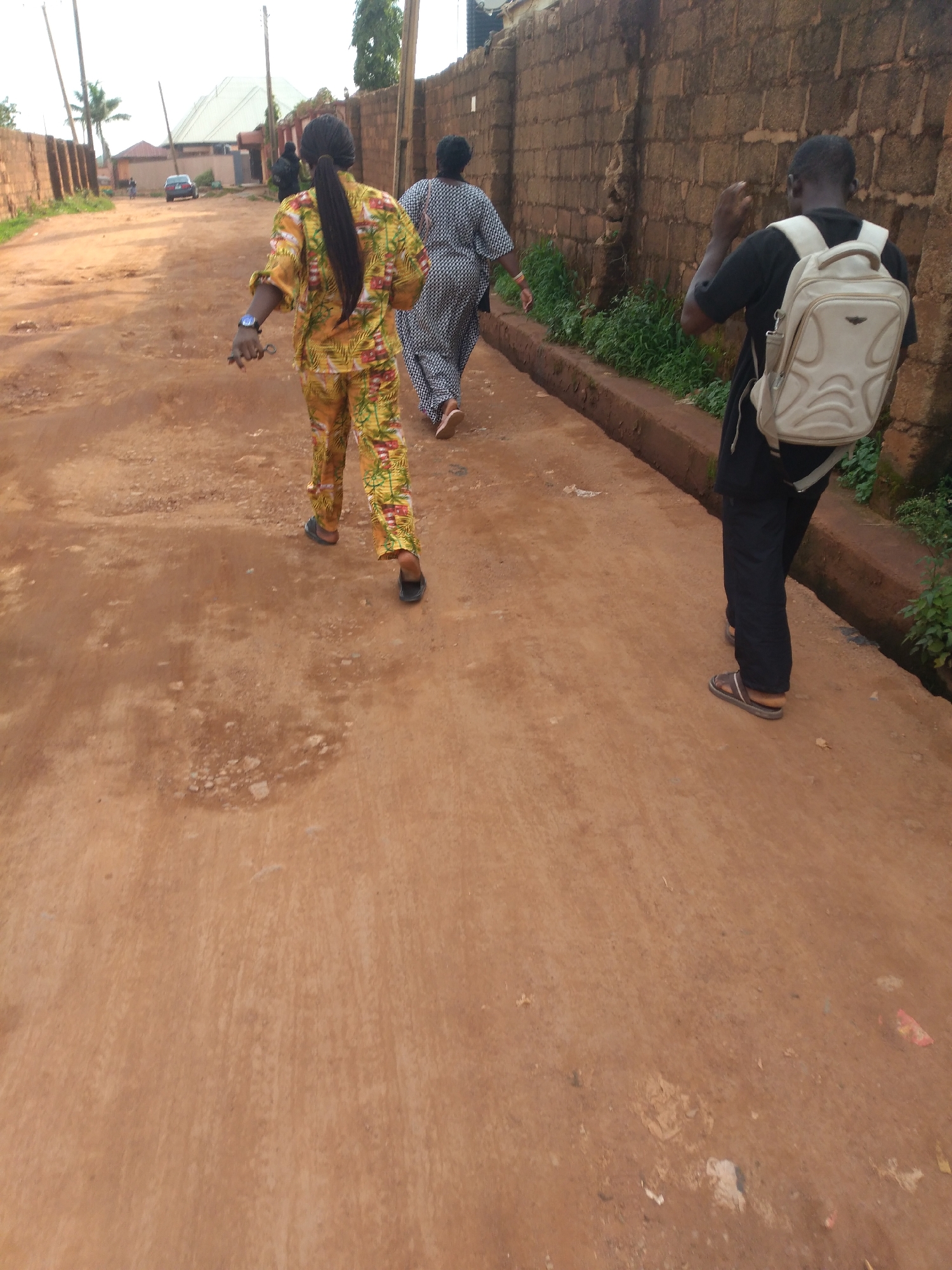 In Jos, Nigeria, three individuals walk down a dusty, unpaved road. On the left, a person wearing a vibrant yellow and green patterned outfit with a long ponytail walks away from the camera. They have a watch on their left wrist and are holding something small and dark in their hand. To their right and further down the road, another person in a black and white patterned dress walks in the same direction. On the far right, a person with a light-colored backpack walks in the same direction, wearing a black t-shirt and black pants. They are holding their right hand up. The road is lined with a low, reddish-brown brick wall on the right, with some vegetation growing along its base. In the background, palm trees, buildings, and a car are visible. The overall impression is of a casual stroll on a warm day in an urban environment.