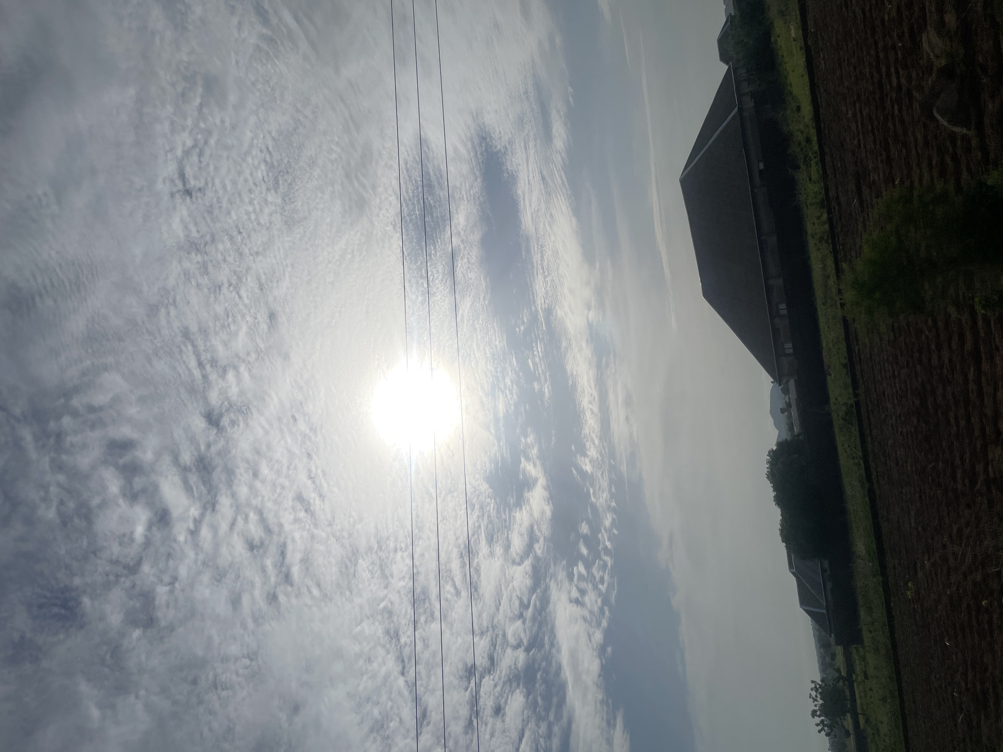 The image captures a view of the city of Jauro Yinu, Nigeria, under a bright, cloudy sky. The sun is visible and appears to be high in the sky, its light diffused by the clouds. Three horizontal power lines stretch across the upper half of the image. In the middle ground, a large, dark-roofed building with a steeply pitched roof dominates the scene. To the left of this building, there is a cluster of trees and another smaller building visible. The foreground shows a field with tilled soil, suggesting agricultural activity. The overall environment appears to be rural or semi-rural. There are no people visible in the image.
