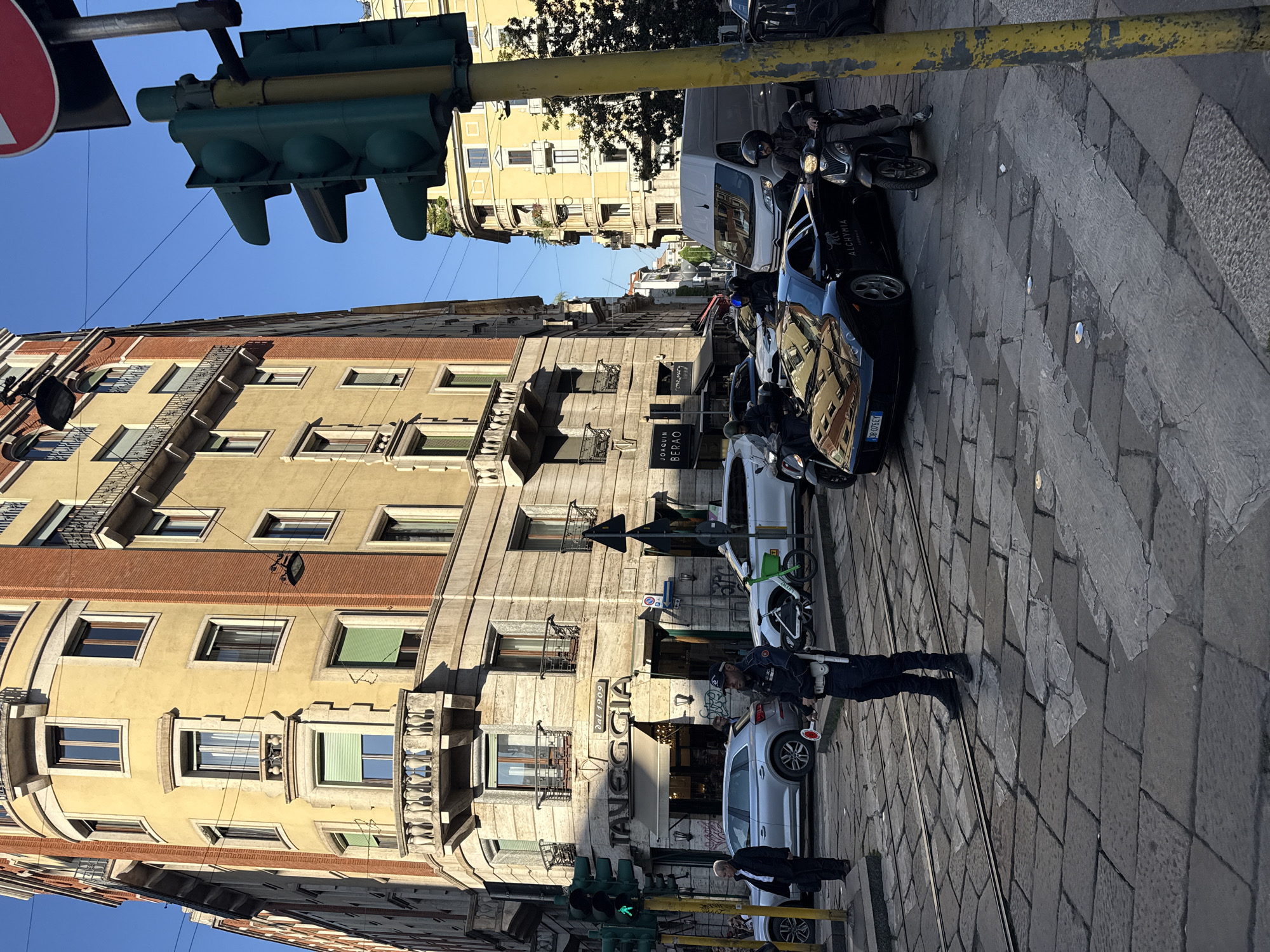 This daytime image captures a bustling street scene in Milan, Italy, under a clear blue sky. The perspective is slightly elevated, looking down a city street lined with multi-story buildings featuring classic European architecture.
On the left, a prominent building with a warm yellow facade, orange-red trim, and ornate balconies dominates the frame. Overhead, a traffic light structure displays a red circular sign with a white bar (indicating 'no entry' or 'do not enter') and a green pedestrian light showing a walking figure.
The street below is paved with irregular grey stone blocks and features tram tracks running through its length. Various vehicles are present, including several parked scooters and motorcycles, along with cars in black, white, and silver. A black car has "ALCHYMIA" visible on its side and a license plate partially reading "DB 076ET."
Several individuals are engaged in street activities. In the midground, a person in a dark uniform, likely a traffic controller or police officer, stands near the tram tracks holding a red and white signaling baton. Another person in a dark suit is also visible nearby.
Notable details include architectural elements like decorative balconies and shutters, bright sunlight casting distinct shadows, and various street signs. Visible text includes "JOAQUIN BERAO" on a building sign, and "TANEGGIA DAL 1909" on a shopfront awning, suggesting a historical establishment. The scene conveys an active urban environment with a blend of historical charm and modern street life.