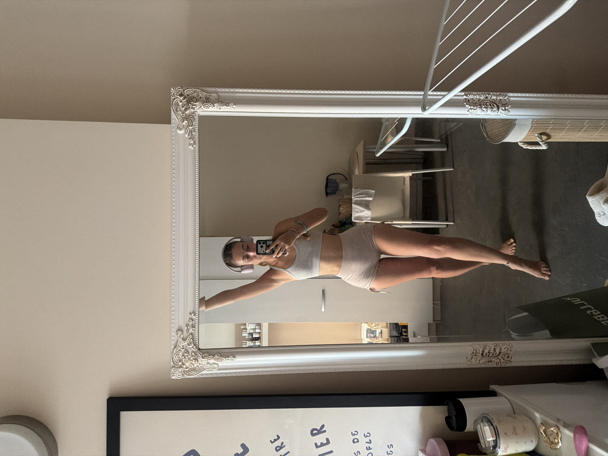 A full-body mirror reflects a young woman standing and taking a selfie. She is wearing a light gray sports bra and light brown shorts, with pink headphones on. She has light brown hair and is looking at her phone, which she is holding in her right hand. Her left arm is outstretched, reaching towards the mirror. Her legs are crossed, and she is barefoot.
The room appears to be a bedroom or dressing area. In the reflection, a drying rack with clothes can be seen in the background, suggesting the room might be used for laundry or clothing storage. There are also shelves with various items, including bottles and containers. The walls are a neutral beige color.
Below the mirror, a framed picture is partially visible, with handwritten text on it. The text includes words like "RKE" and "MER," as well as numbers and symbols. On a surface below the mirror, there are several objects, including what appears to be a candle in a glass jar and other small decorative items.
The lighting suggests it could be daytime, though no windows are clearly visible. The overall atmosphere is casual and personal, with the woman seemingly posing for a photo in her home. The reflection in the mirror provides a clear view of the woman and her surroundings.