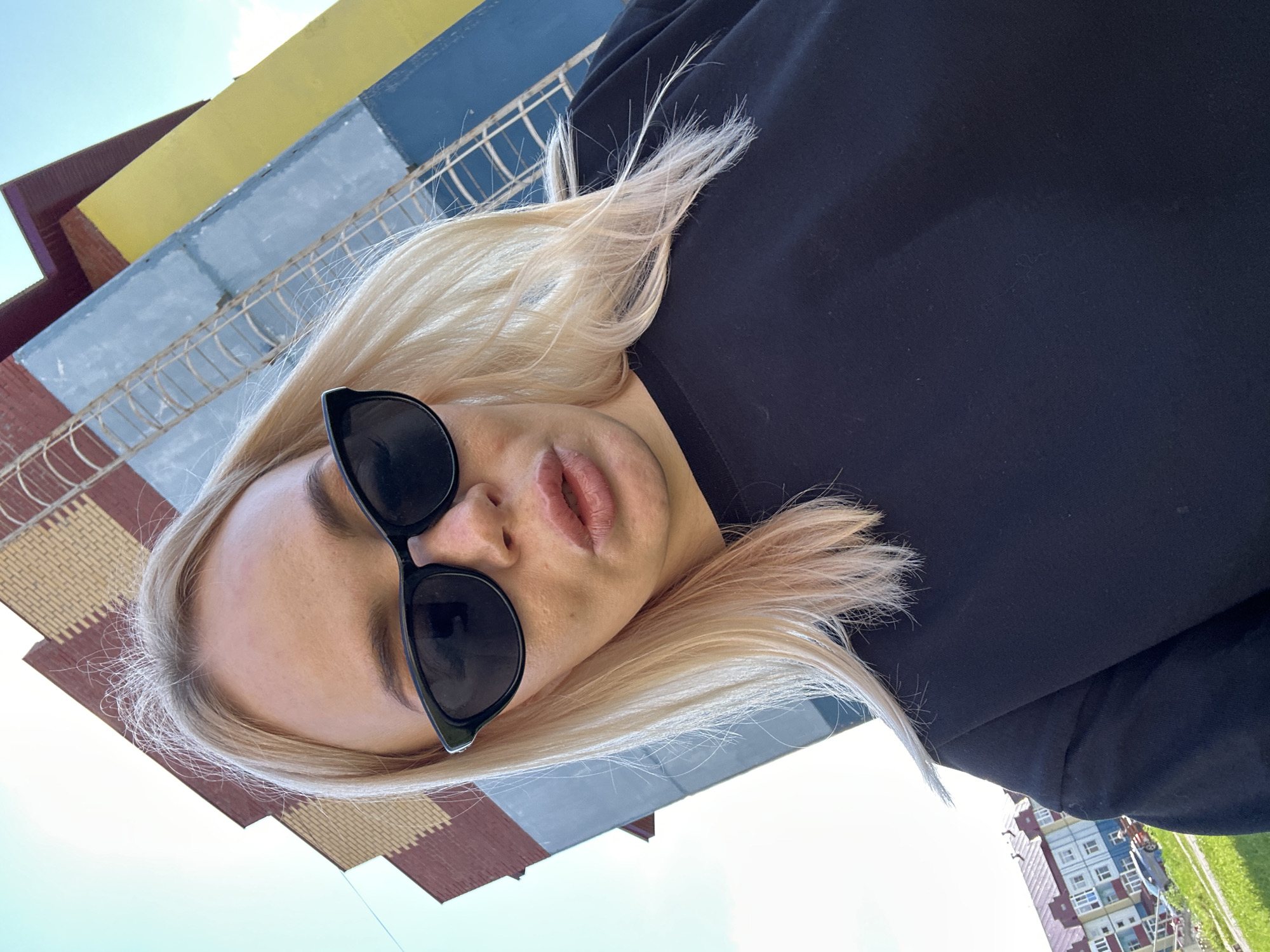 A blond-haired woman wearing black sunglasses and a black sweatshirt is taking a selfie in front of a building. The building is painted in sections of yellow, blue, and tan bricks with a fire escape. In the background, there is a residential building with a blue roof and a small car parked in front. The sky is clear and blue. The woman's mouth is slightly open, and her hair is blowing in the wind.