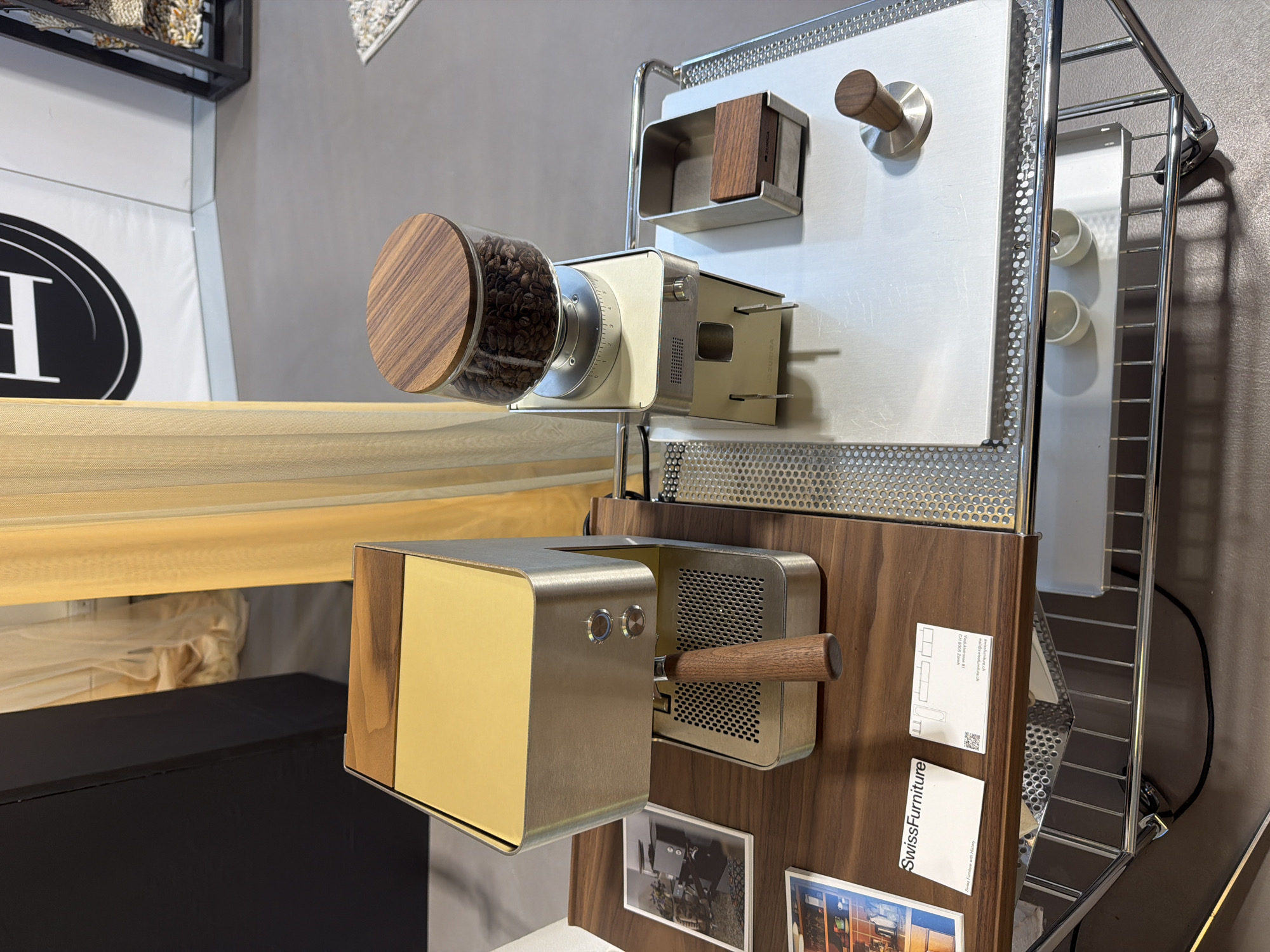 This media file presents an indoor display, likely in a showroom or at an exhibition in Milan, Italy, showcasing a sophisticated coffee preparation setup integrated into a modular furniture system.
The primary subjects are two sleek, modern coffee appliances. On the left, a beige-colored coffee grinder features a clear glass hopper filled with dark roasted coffee beans, topped with a warm wooden lid. A metallic dial with grind settings from 0 to 3 is visible. To its right, a matching beige and brass-toned espresso machine unit is presented, equipped with two circular buttons on its front panel and a wooden handle extending from a perforated metal side, suggesting a portafilter or brewing lever. The brand name "VOLUNGA" is subtly visible on the side of this unit.
These appliances are meticulously integrated into a modular furniture system composed of polished chrome frames, perforated metallic panels, and rich wooden elements. A wooden panel serves as the mounting surface for the espresso machine. Above the grinder, a small wooden block-like container is affixed to a perforated metal sheet, potentially for grounds or accessories. Another standalone wooden knob or handle is mounted on a separate perforated panel higher up, possibly for a coffee tamper. To the right, a chrome wire shelf holds a white tray containing two small white cups.
The background includes a large white fabric banner or display on the left with a partially visible black oval logo featuring a prominent white "H". Below this, a black surface, possibly a table or cabinet, is seen, with a light-colored, translucent fabric draped above it. The backdrop is a neutral gray wall.
Several pieces of text are discernible on white cards attached to a wooden panel. One card clearly states "SwissFurniture" and below it, "Swiss Furniture with History." Another partially visible card includes "ModulMöbelSystem 1," "Vollholzplattenbau AG," "CH 6019 Beromünster," and a QR code, indicating product information.
The scene depicts a static, curated product presentation, emphasizing a blend of modern design, material quality, and functional integration, typical of a design-focused event or retail space. No individuals are present, and the lighting is even, suggesting an artificial indoor setting.