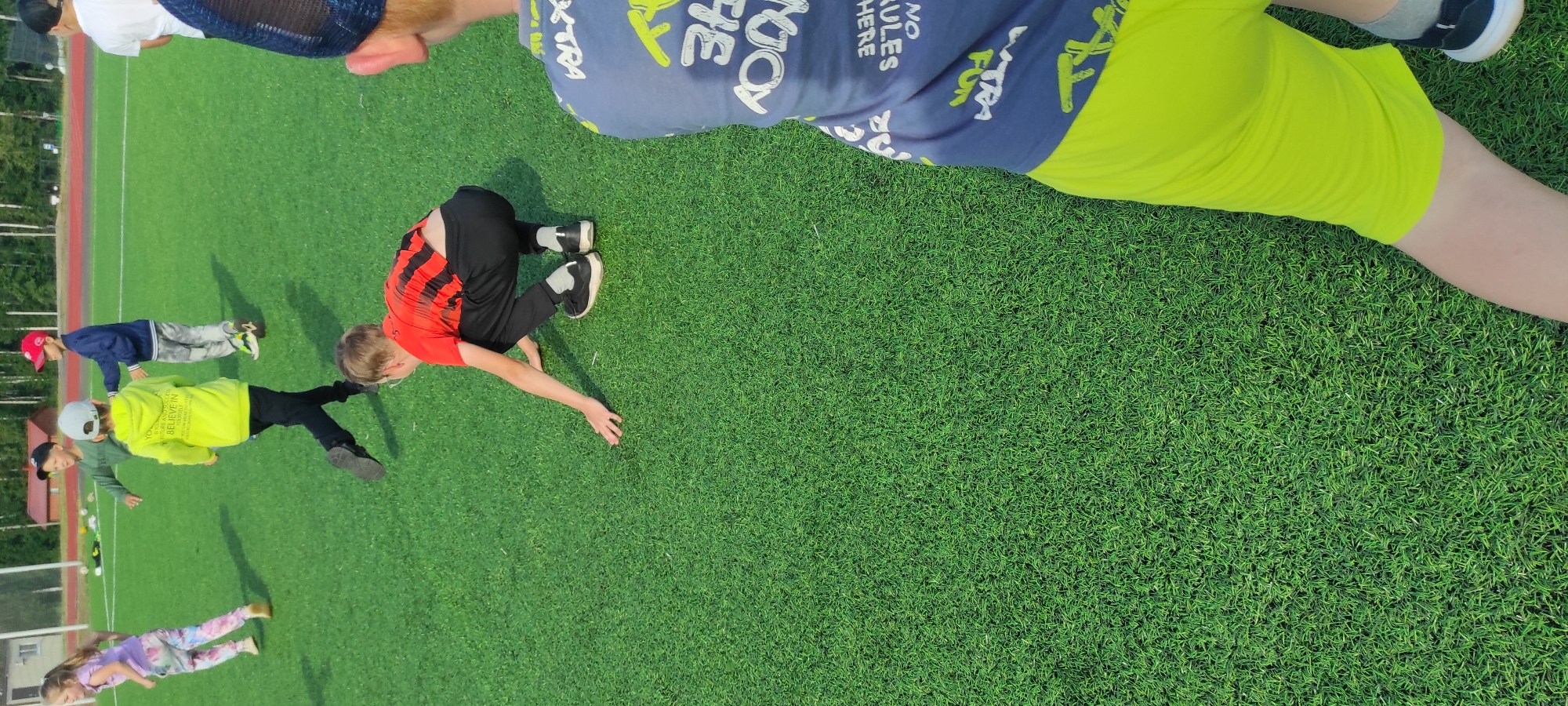 The image depicts a group of children on a bright green artificial turf sports field, likely a soccer field, under daylight conditions. The field appears to be well-maintained. In the background, there are trees and a building, suggesting an outdoor recreational area.
Focusing on the foreground, a boy wearing a red and black striped shirt and black pants is in a squatting position, with his hands reaching towards the ground. Another child, partially visible on the right side of the frame, wears a blue t-shirt with white text and yellow accents, and bright lime green shorts. This child is also on the turf field.
Several other children are visible further in the background. One boy in a bright yellow hooded sweatshirt with text on the back is also in motion, possibly running. Another boy in a dark blue jacket and a red cap is standing. A girl in a purple t-shirt and tie-dye pants is running. The children are all wearing casual sportswear.
The overall impression is one of an outdoor sports activity or gathering involving children on a sunny day. The location is identified as Noviy, Russia, indicating a specific geographical context for this scene of children's recreation.