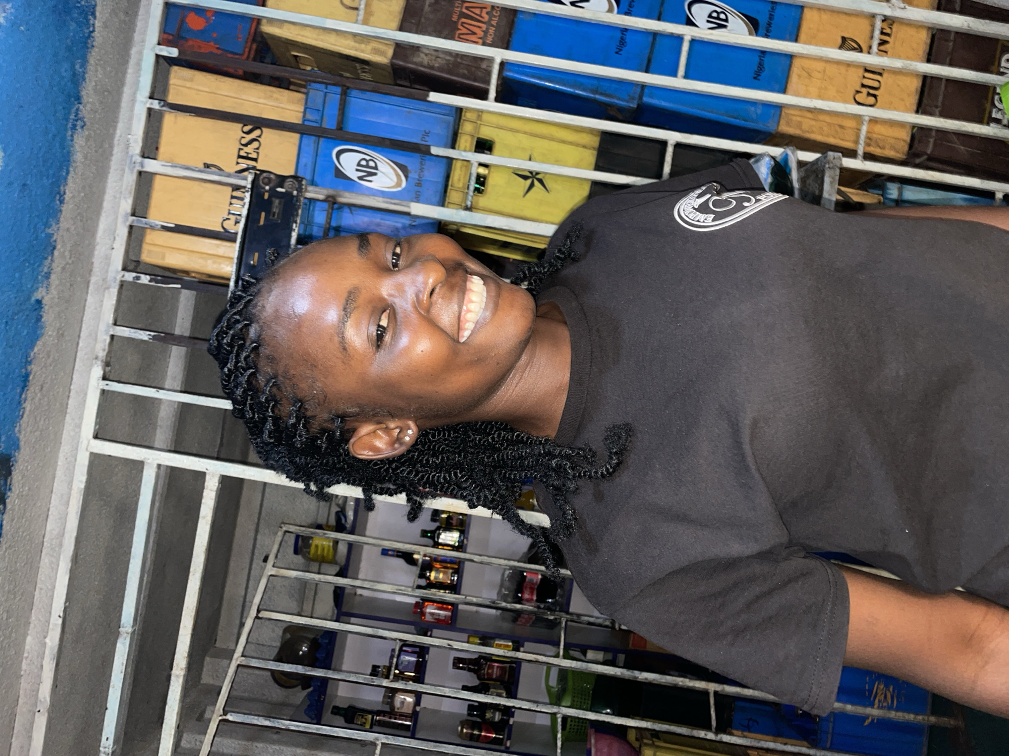 The image captures a smiling young person in what appears to be a beverage retail or storage area in Jos, Nigeria. The individual, with dark skin, is looking directly at the camera with a wide, cheerful smile, revealing white teeth. They have short, dark, tightly coiled braids or twists as a hairstyle and are wearing a dark (likely black) t-shirt with a circular white logo on the right shoulder. A small stud earring is visible in their left ear.
The background is dominated by metal grates and shelving filled with stacked crates and bottles. Many crates are yellow and blue, displaying brand names such as "GUINNESS" and "NB" (Nigerian Breweries Plc). Other text visible on crates includes "MULTIMALT NON ALCOHOLIC." Below these crates, on a lower shelf unit, numerous bottles of various shapes and colors are stored, likely spirits or other packaged beverages.
The scene suggests an indoor commercial setting, possibly a shop, bar, or warehouse, characterized by organized storage of drinks. The lighting appears to be artificial. The subject's expression conveys happiness and amiability.