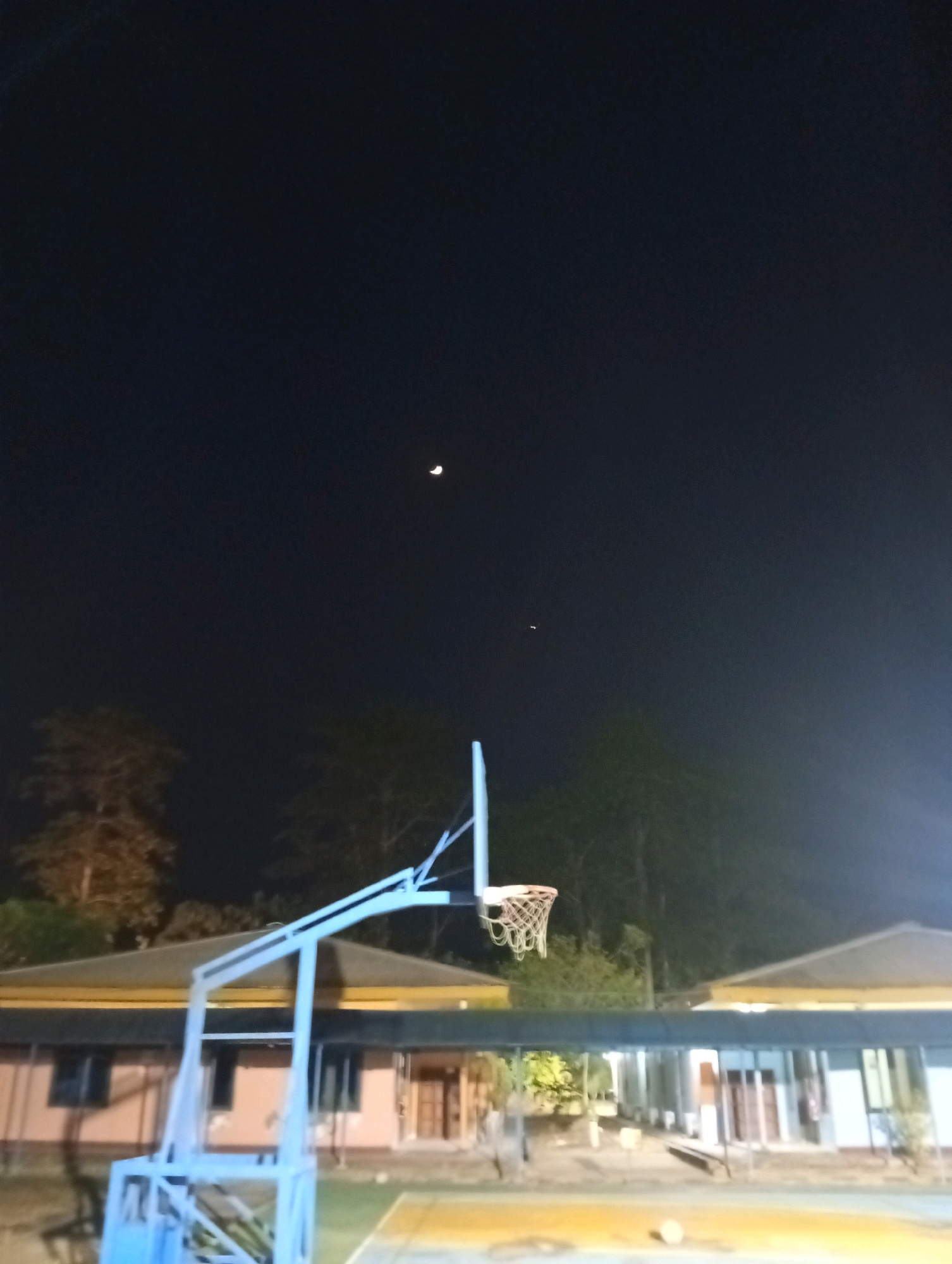 An outdoor basketball court is visible at night in front of a row of buildings with yellow roofs. A basketball hoop with a net is positioned in the foreground. The sky is dark, with a crescent moon and a small bright light, possibly a planet or star, visible. Trees are present in the background. The court surface is visible in the lower portion of the image, with a faint yellow marking. There is a basketball lying on the court. No people are visible in the scene.