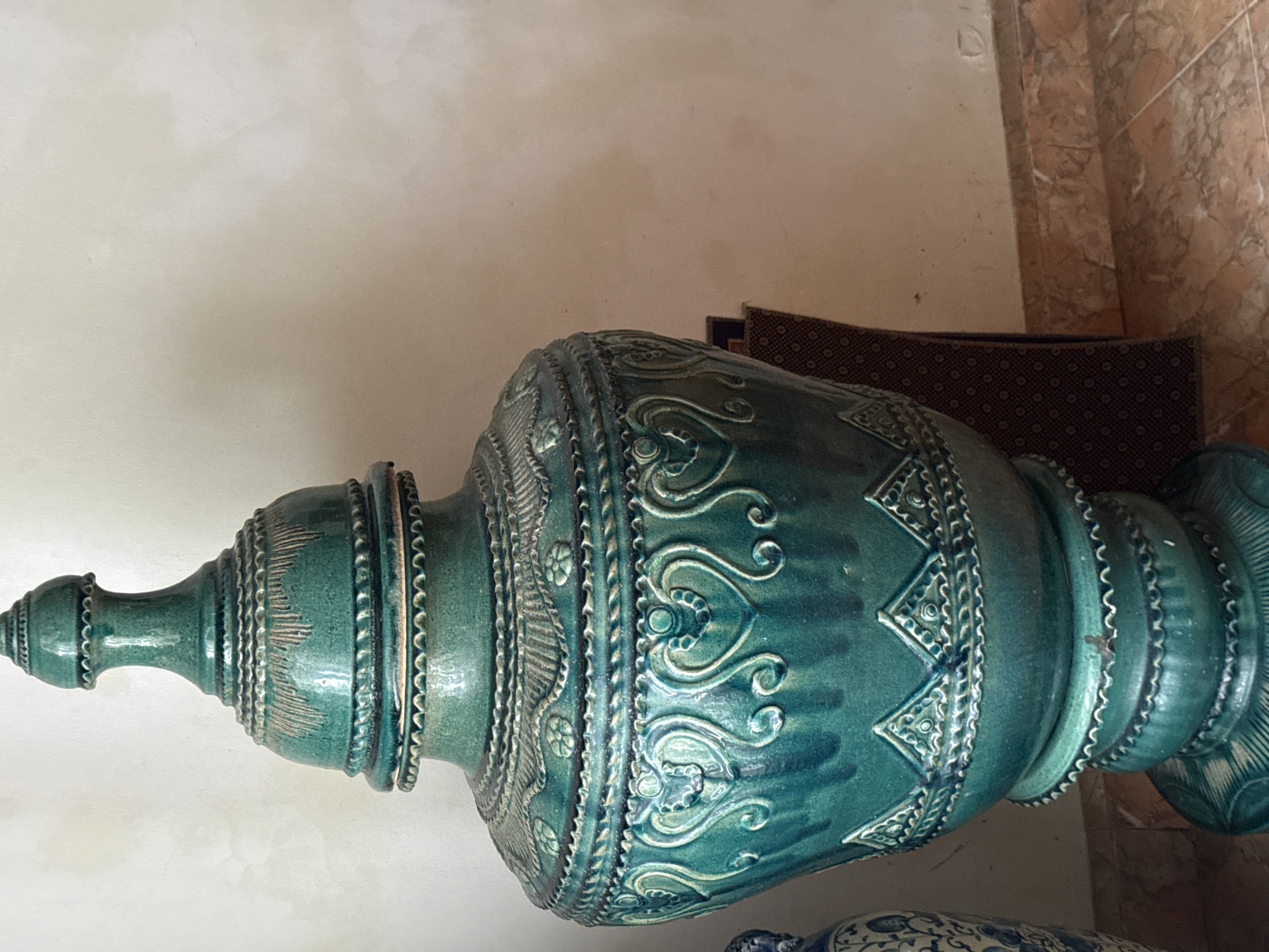 The image depicts a large, ornate, teal-colored ceramic vase with a lid, placed against a light-colored, textured wall. The vase features intricate decorative patterns, including embossed heart shapes, floral motifs, and geometric designs. It rests on a tiered base, also decorated. To the left, a portion of another vase, possibly blue and white, is visible. The floor is tiled with a marbled pattern in shades of orange and brown. A dark patterned object, possibly a piece of furniture or fabric, is partially visible behind the large vase. The scene appears to be indoors, with natural light illuminating the objects. There is no indication of the specific location being Bireuenmeunasahblang, Indonesia; this information is not visually verifiable from the image.