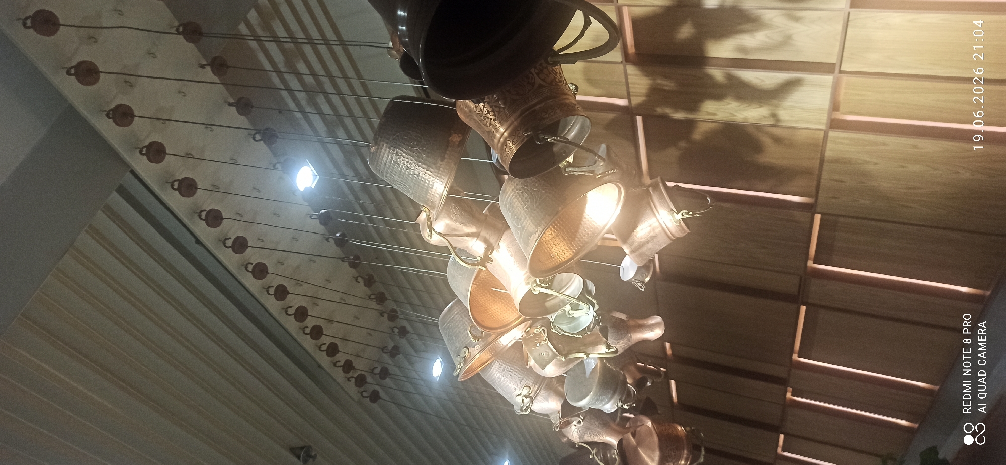 In an interior setting, a collection of copper or brass colored hanging lanterns, shaped like buckets and bells, are suspended from the ceiling. The lanterns feature intricate embossed patterns and are illuminated by overhead lights, casting shadows on a wood-paneled wall in the background. The ceiling appears to be made of light-colored wood planks. The bottom of the image shows a watermark indicating "REDMI NOTE 8 PRO AI QUAD CAMERA" and a date and time stamp of "19.06.2026 21:04." There are no people or animals visible in the scene.