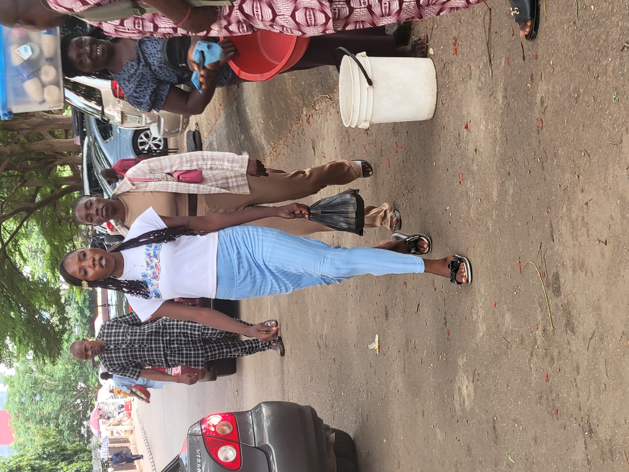 The image captures an outdoor scene in Abuja, Nigeria, depicting several individuals and objects in a street setting.
A young woman with braided hair, wearing a white t-shirt and blue pants, stands out in the foreground. She faces the camera with a neutral expression. Behind her is an older man wearing a black and white checkered shirt. To the left, there are trees and buildings that suggest the location is in a city. To the right, there are several cars parked on the street.
Several other individuals can be seen further up the street, including one person holding a red bucket, another holding a bucket, and another person holding a bag.
The ground is paved and shows signs of wear.