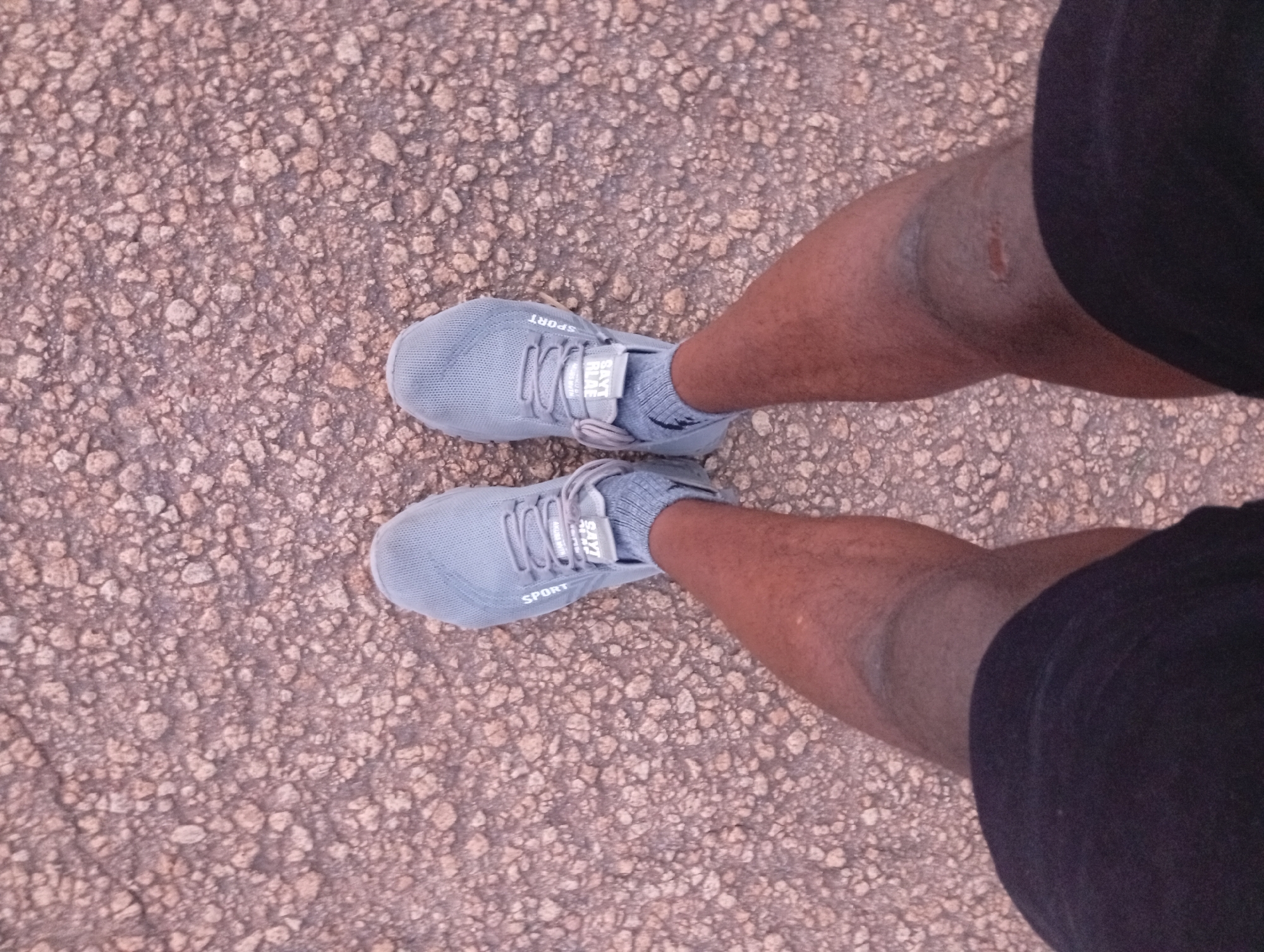 The image displays a top-down view of a person's legs wearing grey athletic shoes and dark shorts, standing on a textured ground surface composed of small, light-colored pebbles and dirt. The shoes have white laces and a grey strap with white lettering that reads "SPORT". One shoe has a tag with white text. The ground appears to be an outdoor path or unpaved area, consistent with a location in the city of Jos, Nigeria, known for its rocky terrain and extensive use of such materials for pathways and landscaping. There are no other people or significant objects visible in the frame.