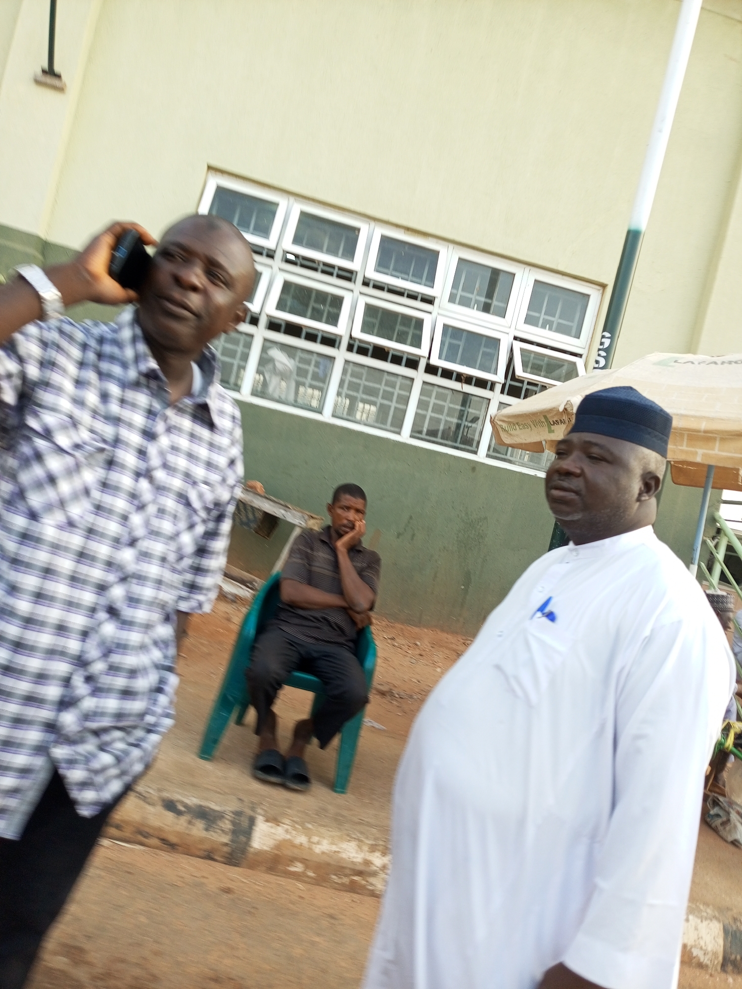 In Gombe, Nigeria, two men stand outside, with a third man seated in a chair in the background. The man on the left, wearing a checkered shirt, is holding a mobile phone to his ear. To the right, a man in a white robe and a dark cap looks to his left. The seated man in the background, wearing a dark shirt and pants, leans forward with his hand on his chin. The environment appears to be an outdoor area with a building that has several windows in the background, and an umbrella is visible on the right side of the frame. The ground is a mix of dirt and pavement.