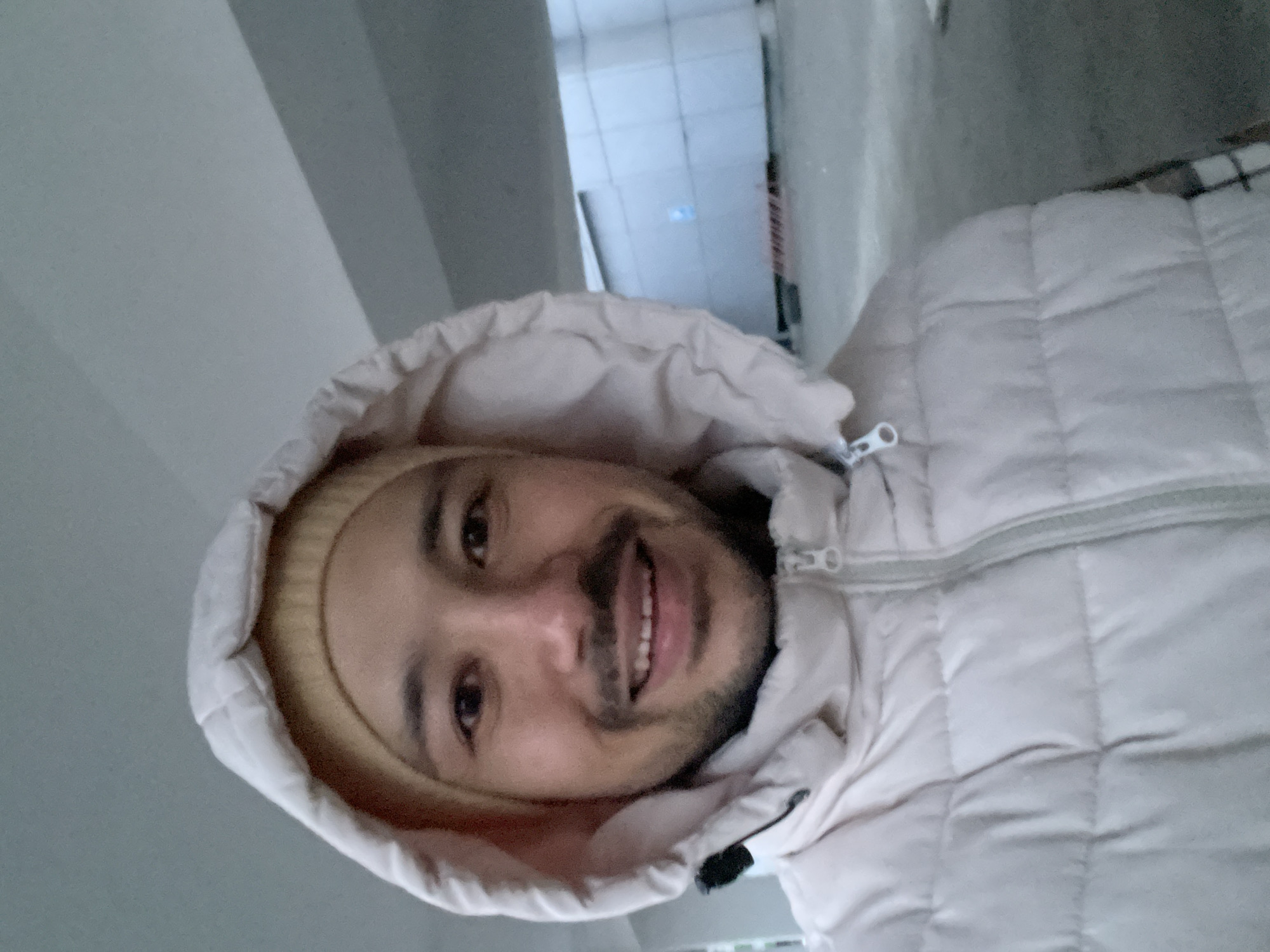 A man with a beard and a beige beanie under the hood of a quilted, light beige puffer jacket smiles at the camera. He appears to be in an indoor corridor, possibly in a building in Izhevsk, Russia, judging by the tile flooring and the general architectural style.