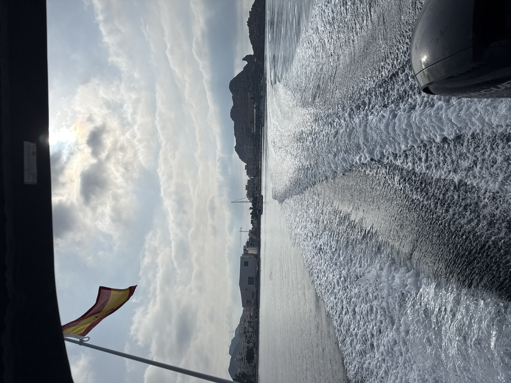 The image shows a view from the back of a boat moving through water, leaving a foamy wake. A Spanish flag flies from a mast on the left side of the frame. In the distance, the coastline of Águilas, Spain, is visible, featuring buildings, hills, and a rocky outcropping. The sky is partly cloudy with the sun breaking through. The lower right corner of the image shows the back of an outboard motor.
