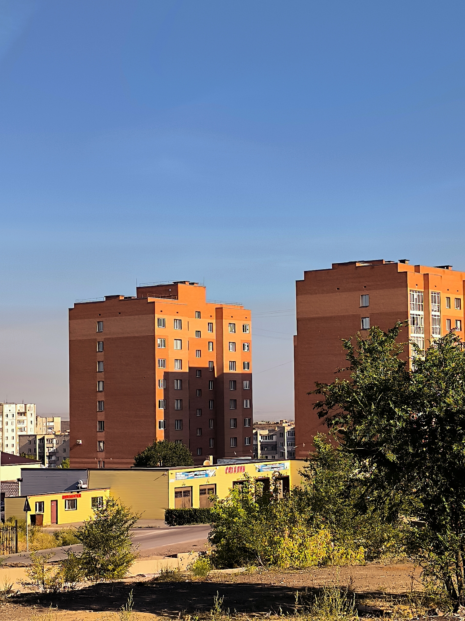 The image depicts a daytime outdoor scene in the city of Qarağandı, Kazakhstan, featuring several buildings and vegetation under a clear blue sky. In the foreground, there is dry, unkempt grass and some dirt. A road or paved area is visible in the middle ground, with a low-lying yellow building situated behind it. This building has several windows and doorways and appears to have signage on its facade, some of which is in Cyrillic script. To the left of this yellow building, a gate and fence can be seen.
Dominating the background are two tall, modern apartment buildings constructed from orange-red brick. The building on the left is somewhat darker and features evenly spaced windows. The building on the right is brighter orange and has more complex window arrangements, including some with balconies or enclosed sunrooms. In the far background, other buildings, including a lighter-colored apartment complex, are visible, suggesting an urban or suburban environment. Lush green trees are present in the right and left mid-ground, partially obscuring the lower sections of the apartment buildings and adding a natural element to the scene. The lighting suggests it might be late afternoon or early morning, with shadows cast on the buildings. There are no people visible in the image.