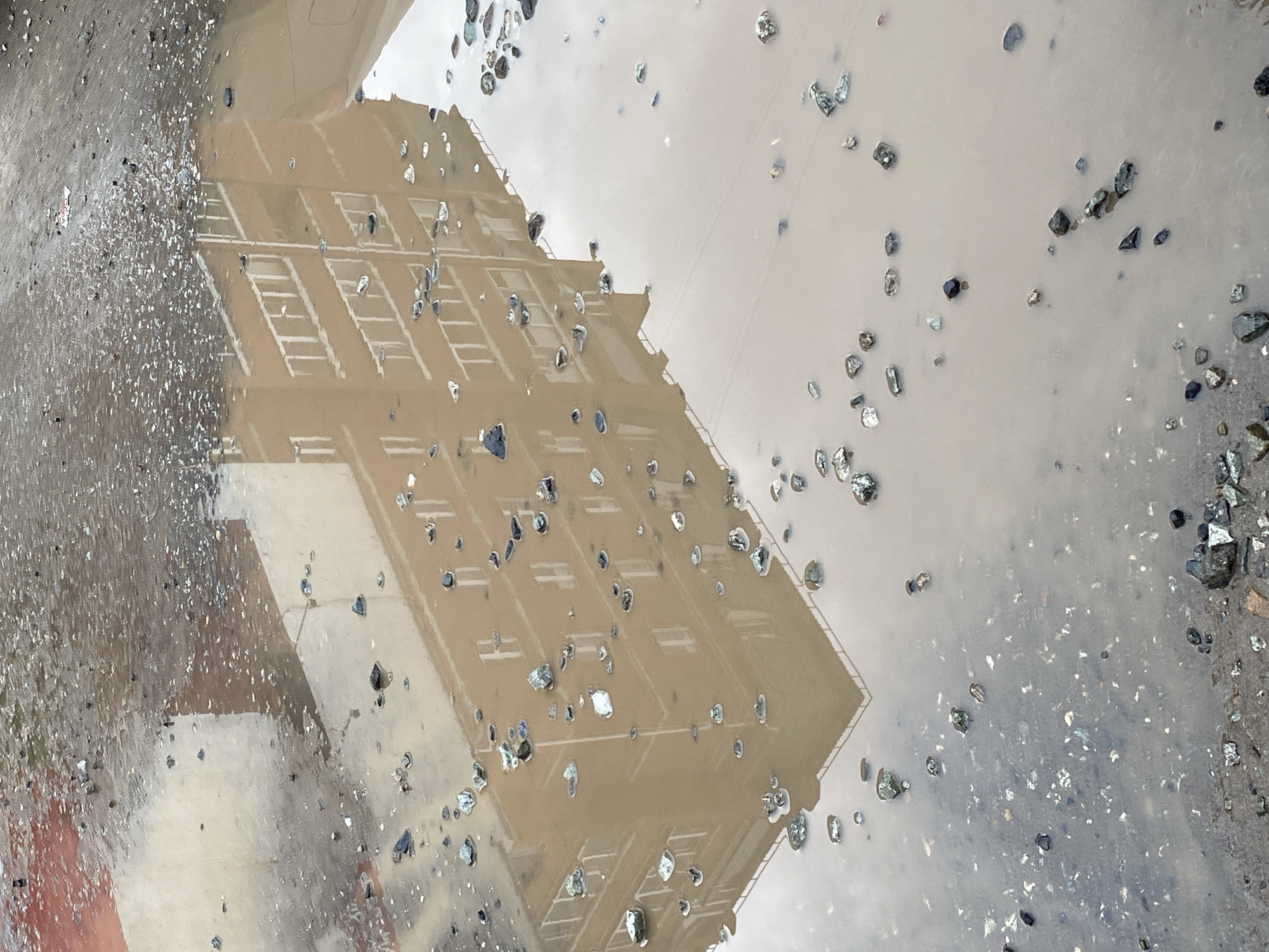 The image depicts a reflection of a building in a puddle. Small stones and debris are scattered around the puddle. The building is multi-story with a light-colored facade and evenly spaced windows and fire escape ladders. The setting appears to be an urban area in Chaikovsky, Russia. The environment suggests recent rainfall. There is no visible people.