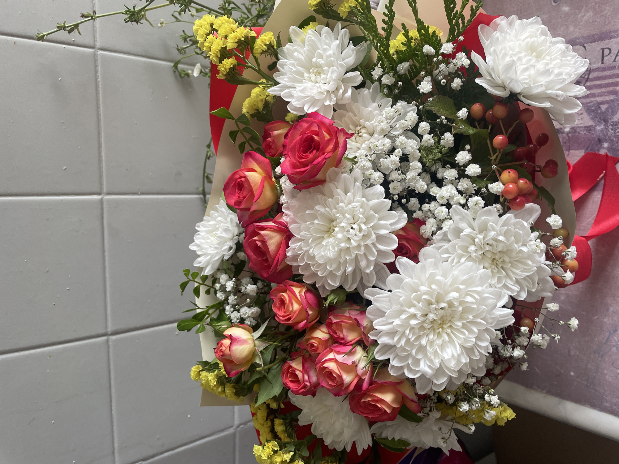 A bouquet of flowers is displayed against a tiled wall. The bouquet features a mix of white chrysanthemums, pink and yellow roses, yellow statice, baby's breath, and red berries. The flowers are arranged in a light brown paper wrap, tied with a red ribbon. The background wall is tiled with square, light gray tiles. The overall scene is a close-up shot of the floral arrangement. There is no information about the city of Brest, Belarus or any people in the image.