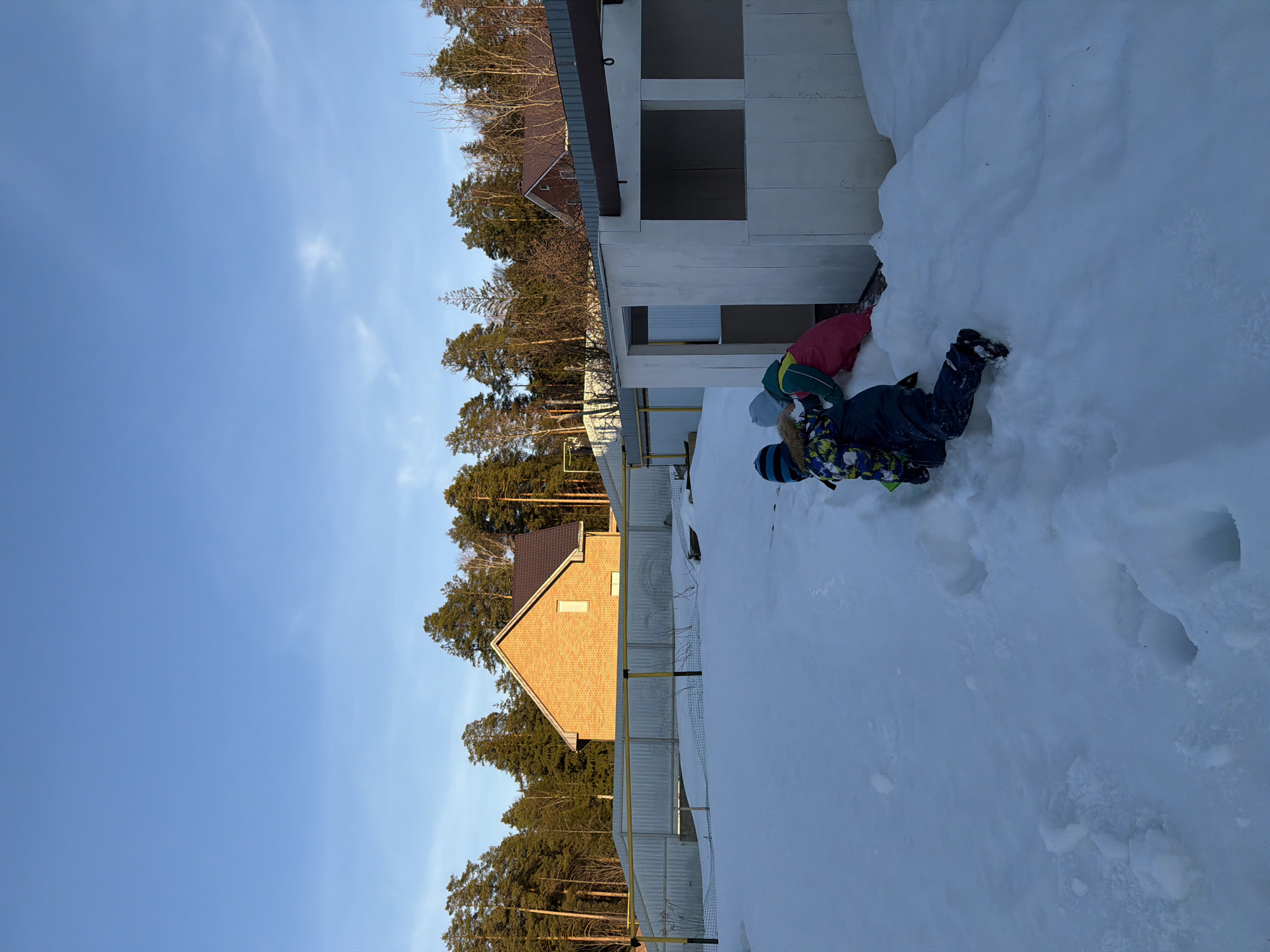Two children are playing on a large snowdrift next to a building under construction. The building is a light gray, two-story structure with rectangular window openings lacking glass. The snowdrift appears to be piled high against the side of the building, almost reaching the second-story windows.
One child is wearing a blue and black snowsuit with a pattern and a blue and black striped hat. The other child is wearing a red jacket, and a green and blue hat, partially hidden. Both children are partially submerged in the snow.
In the background, there are several buildings, including a yellow brick house with a brown roof. There is also a gray metal fence, running parallel to the snowy area where the children are playing. Tall pine trees form a backdrop to the buildings, and a clear blue sky is visible above.
The scene is possibly located in the city of Chaikovsky, Russia.