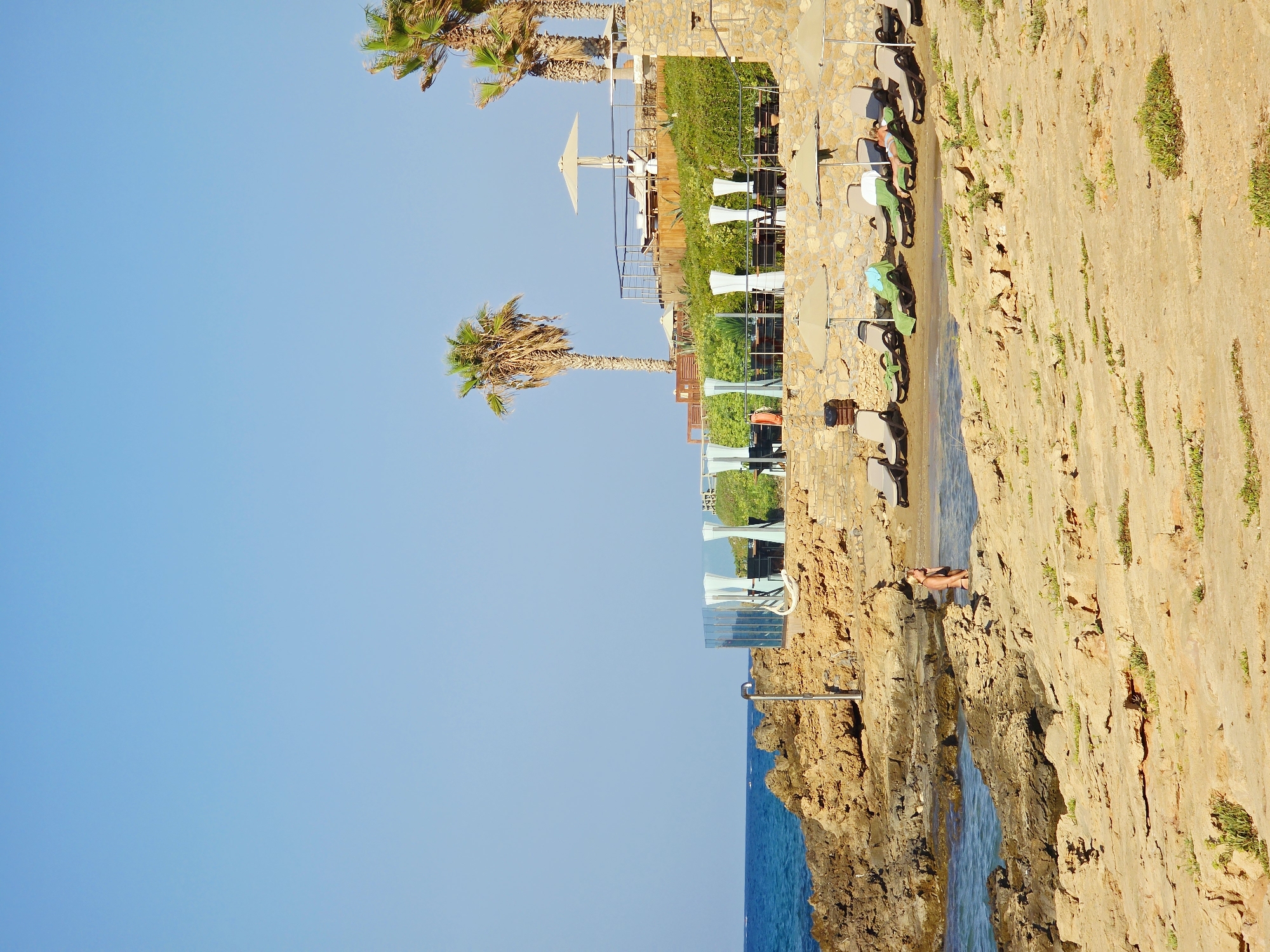 The image shows a rocky coastline with a beach and several sun loungers set up. A woman in a bikini is standing in the shallow water near the rocks. In the background, there is a structure with glass walls and white curtains, possibly a restaurant or bar, with palm trees visible. The sky is clear and blue. The location is the beach area of Malia Municipal Unit, Greece.