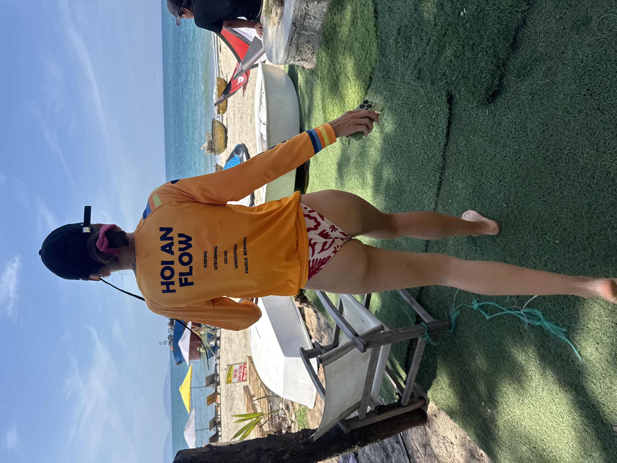 A person stands on artificial turf at a beach in Đà Nẵng, Vietnam. The individual wears a black baseball cap, a long-sleeved orange shirt with “HOI AN FLOW” printed on the back, and red and white patterned swim bottoms. They hold a cell phone in their right hand. The background features a beach with lounge chairs, umbrellas, boats, and the ocean.