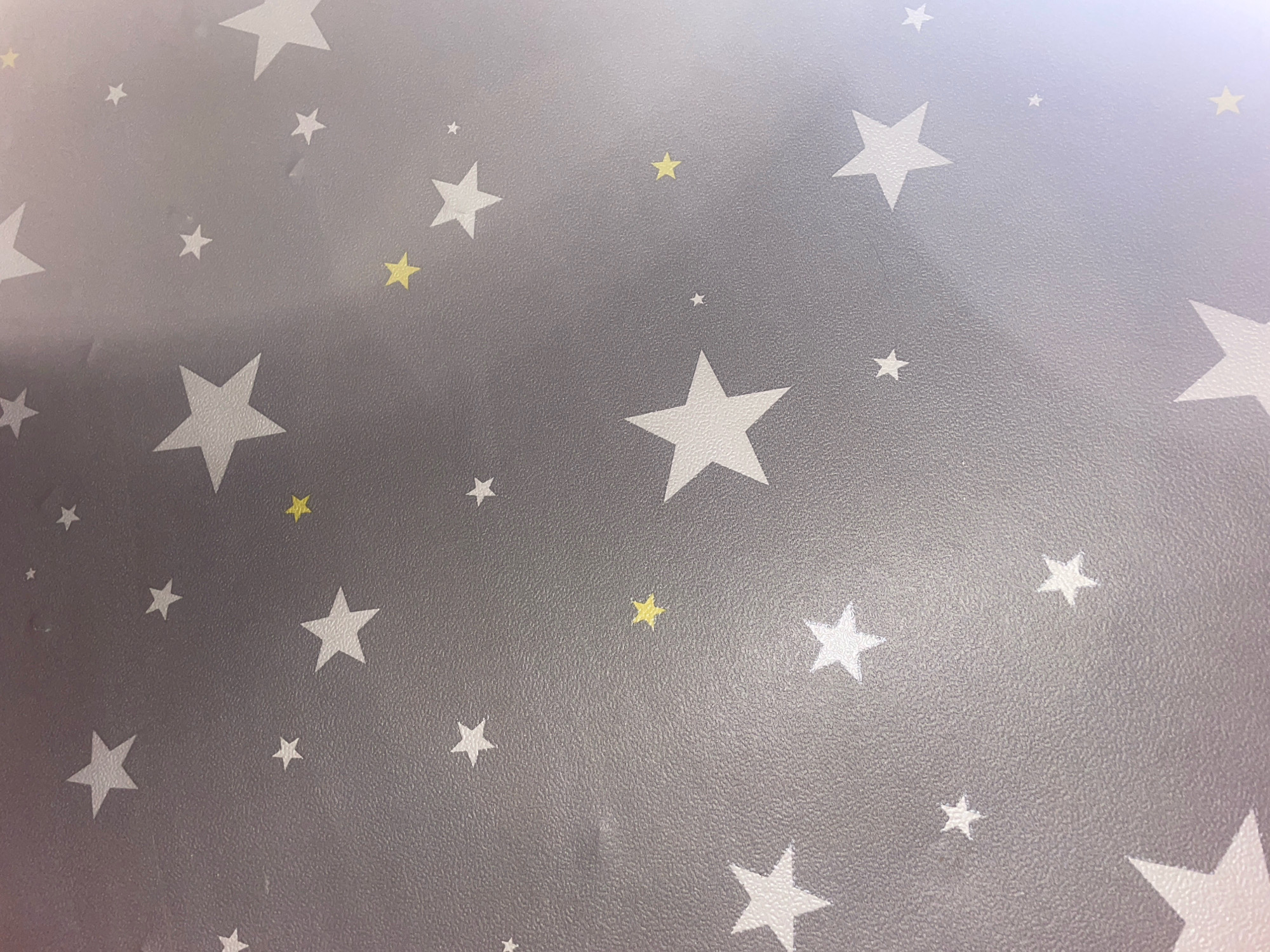 The image displays a close-up view of a grey surface adorned with a repeating pattern of white and yellow stars. The stars vary in size and orientation, creating a whimsical and celestial aesthetic. The surface itself appears to have a slightly textured finish, possibly indicating wallpaper or a similar material. The lighting suggests an indoor setting, possibly a child's room or nursery. The image does not depict any people, actions, or interactions. There are no identifiable landmarks or elements that would place it specifically in the city of Марʼіна Горка, Belarus.