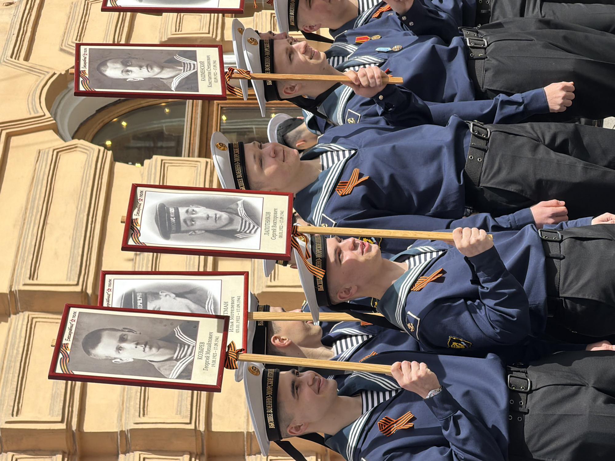 A group of young men in naval uniforms participate in a "Immortal Regiment" march. The marchers are lined up in an urban setting, likely a city street in Saint Petersburg, Russia, as evidenced by the architecture and details of the naval uniforms. Each marcher carries a framed portrait attached to a wooden stick. The portraits appear to be black and white photographs of deceased veterans or relatives, likely participants in World War II, along with their names, ranks, and dates of birth and death. The frames on the portraits feature a ribbon that looks like the Ribbon of Saint George. The naval uniforms consist of dark blue jackets, black trousers with belts, and white sailor hats with the words "ВЫСШЕЕ ВОЕННО-МОРСКОЕ ИНЖ", meaning "Higher Naval Engineering." Some uniforms display a medal or ribbon on the chest. The architecture in the background features a light-colored building with classical elements, including arched windows and decorative moldings. The photograph is taken during the daytime, with natural light illuminating the scene. The marchers look in the same direction, as they are marching.