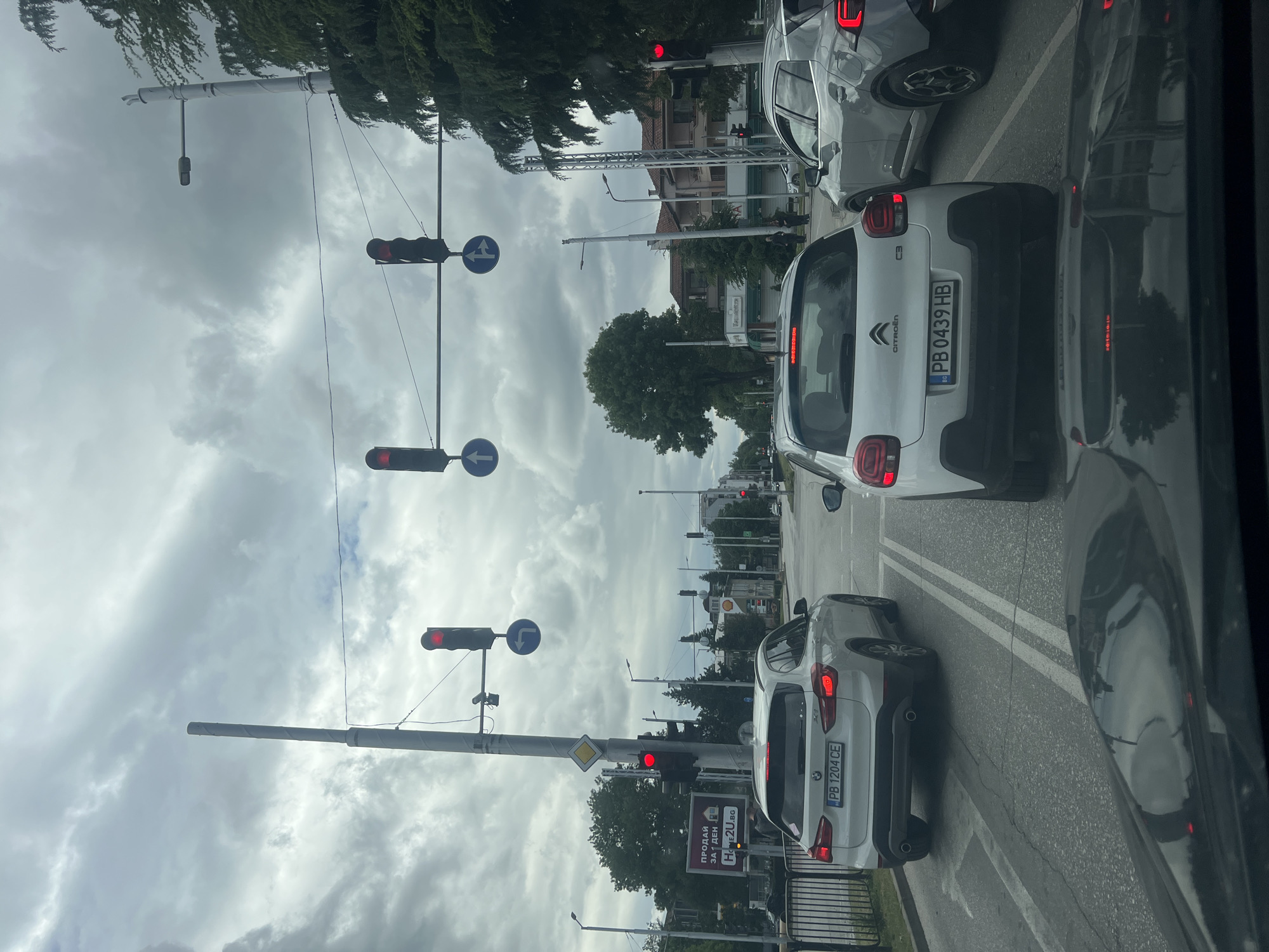The image captures a street scene in Plovdiv, Bulgaria, with a traffic jam at a traffic light. The sky is overcast.
Several cars are visible, including a white Citroën C3 with license plate "PB 0439 HB" and a white BMW X1 with license plate "PB 1204 CE". The traffic lights are red.
Street signs and poles are visible. An advertisement sign reading "ПРОДАЙ ЗА 1 ДЕН HOME2U.BG" is present.