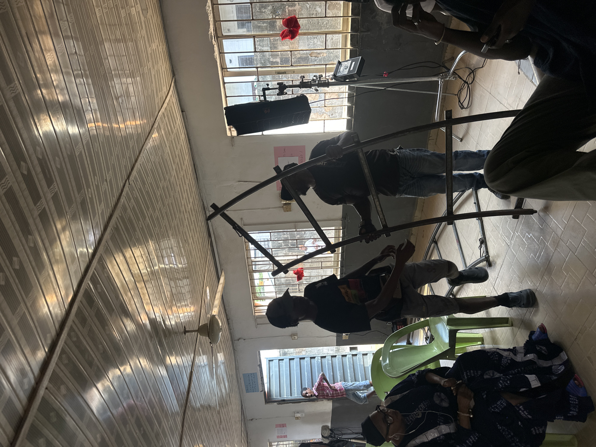 This image captures an interior scene that appears to be a film or video production set in Lagos, Nigeria, illuminated by bright daylight. The room features a distinctive corrugated metal ceiling with reflective patterns and a light-colored tiled floor.
Several individuals are present, engaged in various activities. In the center, a multi-bar metal frame, likely scaffolding or a rig for equipment, is being set up or adjusted by at least two people. One person, wearing a dark t-shirt and jeans, is seen actively working with the frame. Another individual in a dark top and lighter shorts is also interacting with the structure. In the foreground, positioned to the lower right (in the original orientation), a person is resting on a green plastic chair, covered with a patterned cloth, wearing glasses and earphones.
Near the windows, which are fitted with security bars, professional lighting equipment is visible. A large black light fixture is mounted on a stand, facing into the room, alongside a smaller monitor or light. A piece of red fabric is tied to the window bars. Various cables lie on the floor, consistent with a production environment. Through an open doorway in the background, a person in a plaid shirt and shorts is standing outside. A white ceiling fan is mounted on the corrugated ceiling.
Bright sunlight streams through the windows, indicating it is daytime. A pink note taped to the wall has visible text: "15 min is done" and "SAGADO." The overall scene depicts a practical workspace for media production, likely in preparation or during a break.