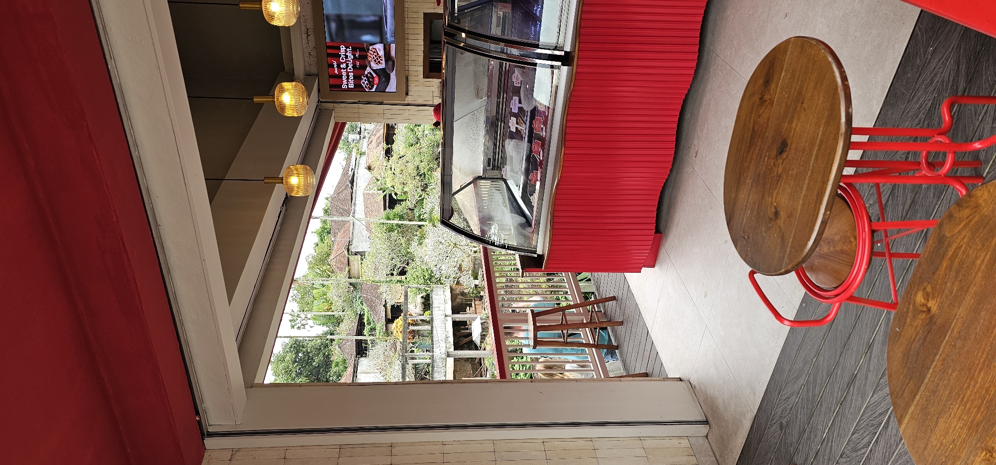 The image shows an interior space, possibly a cafe, located in Ubud District, Indonesia, as indicated by the surrounding architecture and vegetation visible through a large window. The view from the window includes residential buildings and lush greenery, characteristic of the area.
Inside, the cafe features modern furnishings, including round wooden tables with red metal frames and a red display case likely for food items. A flat-screen display shows an advertisement for treats. Three light fixtures hang from the ceiling. The interior design elements such as the red color scheme and the combination of wood and metal suggest a contemporary style.
Outside the window is a view of roofs of houses and the forest.