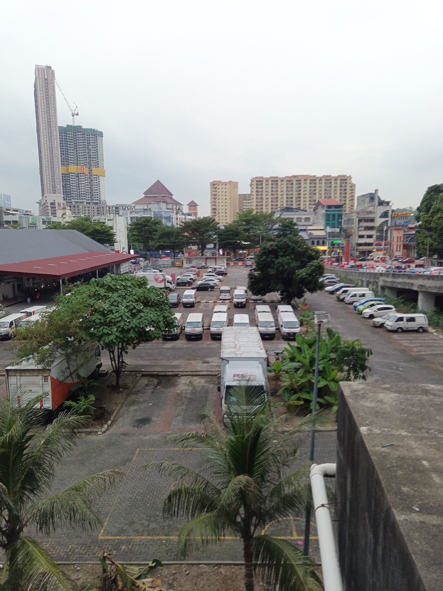 This is an eye-level, medium shot photograph of a parking lot in Kuala Lumpur, Malaysia, on an overcast day. The scene depicts a busy urban environment with various vehicles, buildings, and greenery.
In the foreground, palm trees with fronds frame the lower portion of the image. A concrete wall occupies the right edge of the frame, with a white pipe running along its surface. The parking lot itself is paved with interlocking bricks and marked with yellow lines, indicating parking spaces. Several vehicles are parked here, including white delivery vans and trucks.
Further into the background, a large green tree stands prominently. Beyond the parking lot, a road is visible, lined with parked cars and a bridge overhead. In the distance, a dense collection of buildings of varying heights and architectural styles dominates the skyline. Notable structures include a very tall, slender skyscraper under construction with a crane attached, a building with a distinctive red, pyramidal roof, and several other multi-story buildings. The overall impression is one of a bustling urban center with ongoing development.