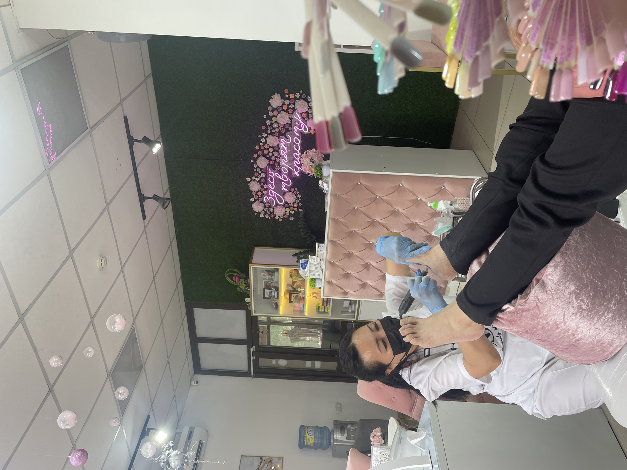 In a beauty salon located in Almaty, Kazakhstan, a female technician wearing a white uniform and a black face mask is performing a pedicure on a client's foot. The technician, with dark hair pulled back, is using an electric nail file to work on the client's toenails. The client, dressed in black pants, is seated with their feet resting on a pink cushion. The salon interior features a green accent wall with a neon pink sign that reads "Здесь творят красоту" (Beauty is created here), surrounded by floral motifs. The room is decorated with hanging decorative balls, a pink tufted reception desk, a display shelf with various products, and nail polish sample sticks arranged in the foreground.